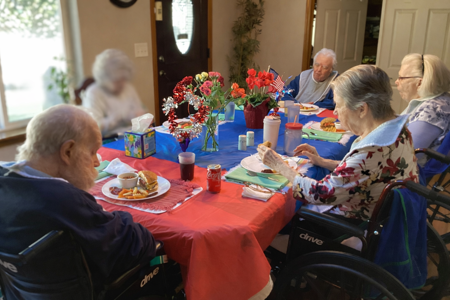 Four elderly people in wheelchairs eating a meal together at a dining table decorated with red and blue tablecloths and floral centerpieces.