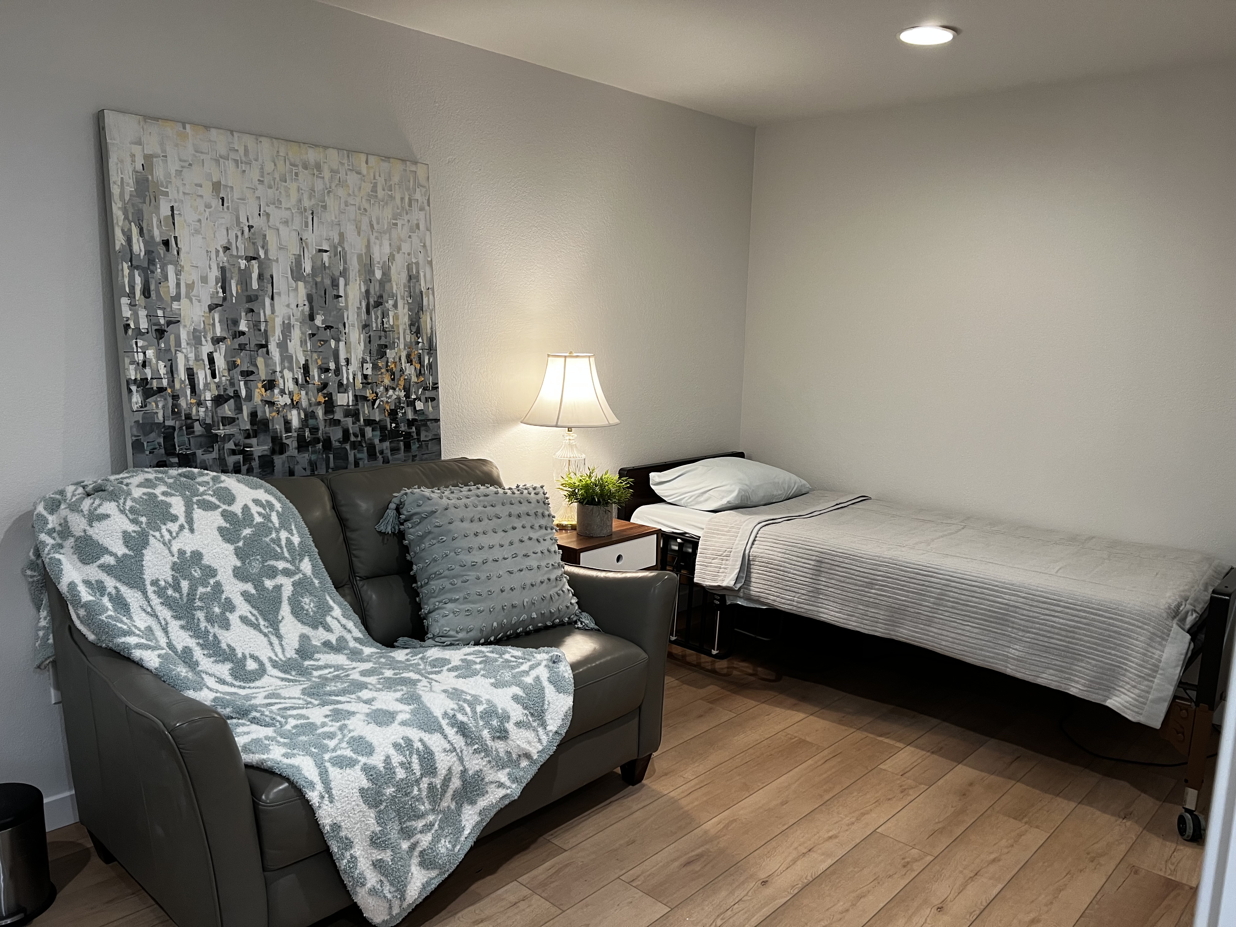 Cozy bedroom with a single bed covered in light gray bedding, a dark leather armchair draped with a floral blanket, a textured pillow, and a nightstand with a lamp and small plant.