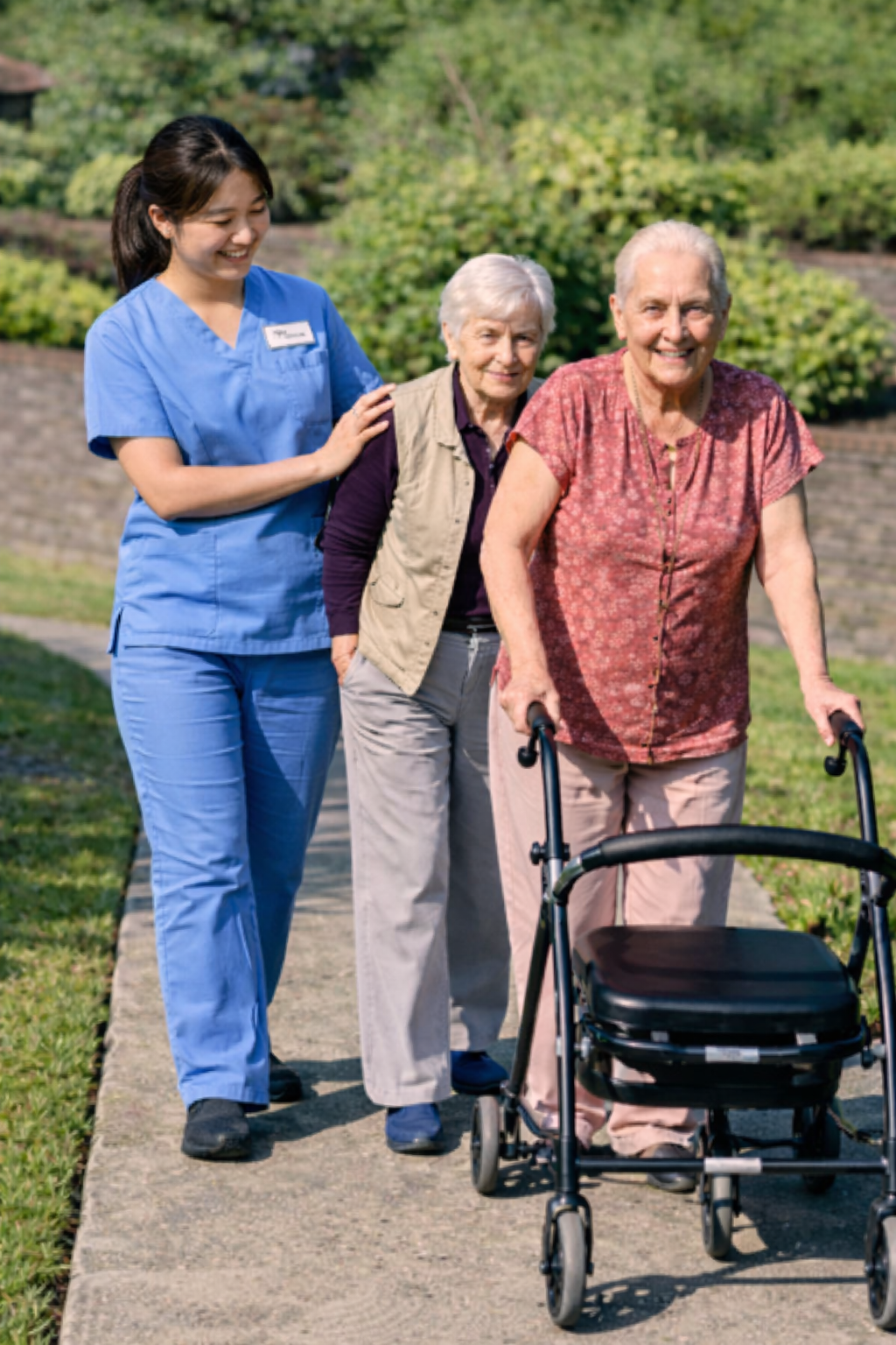 A nurse in blue scrubs smiling and walking outdoors with two elderly women, one using a walker.
