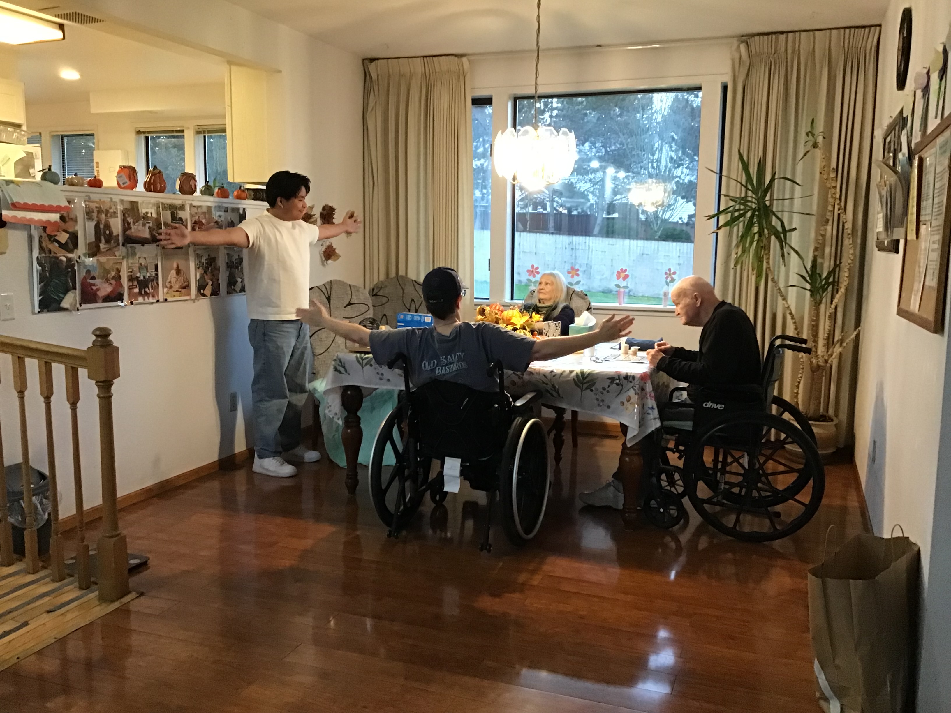 Three people, two in wheelchairs and one standing, with arms outstretched in a warmly lit dining room with photos on the wall and a large window.