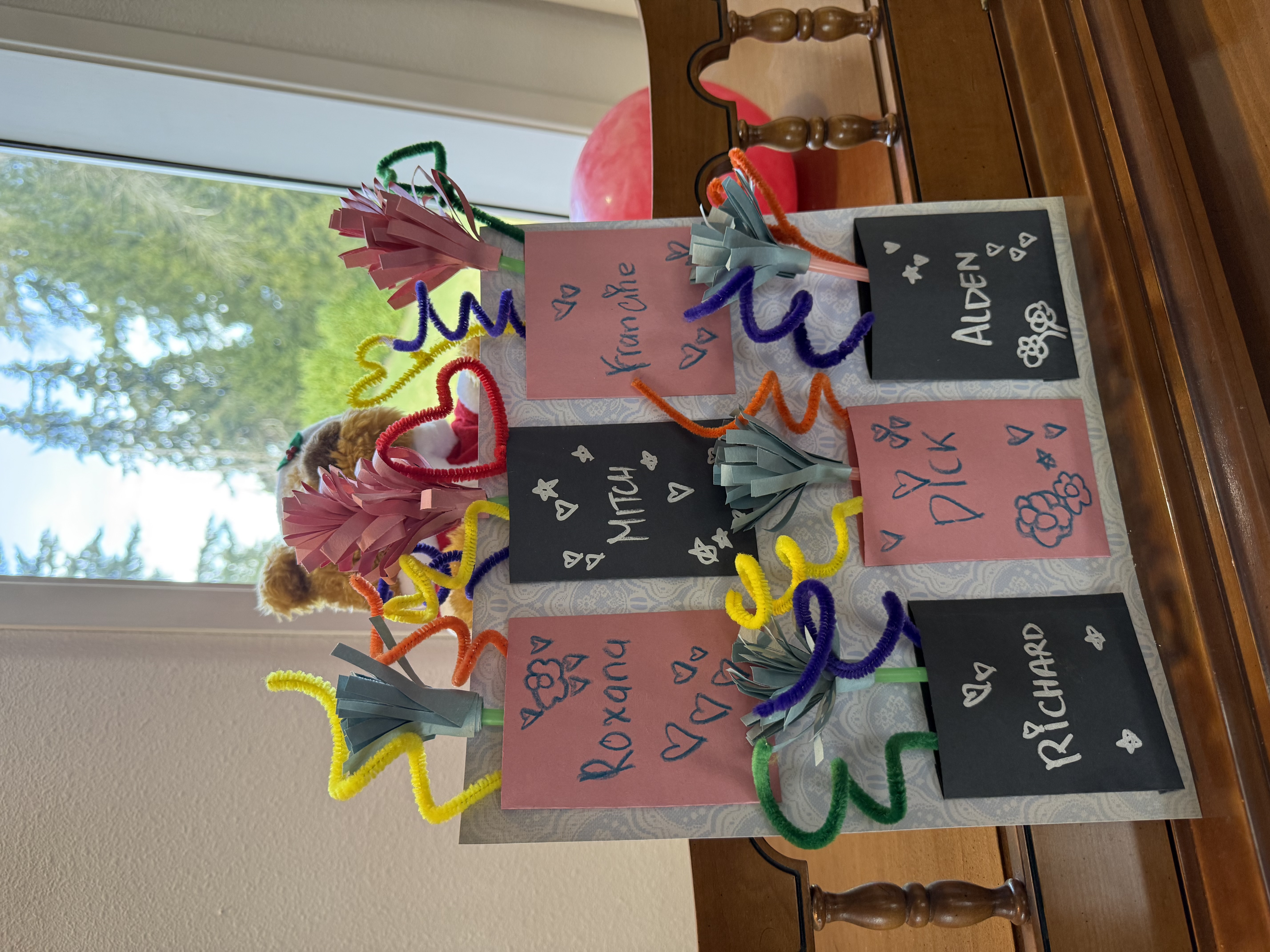 Handmade paper flowers with colorful pipe cleaner stems placed in black and pink paper holders labeled with names Roxana, Mitch, Francine, Richard, Dick, and Alden, on a wooden surface near a window.