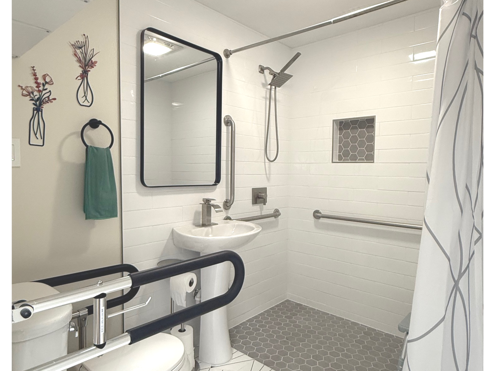 Accessible bathroom with white tiled shower, gray hexagonal floor tiles, grab bars, pedestal sink, black-framed mirror, and green towel on a ring.