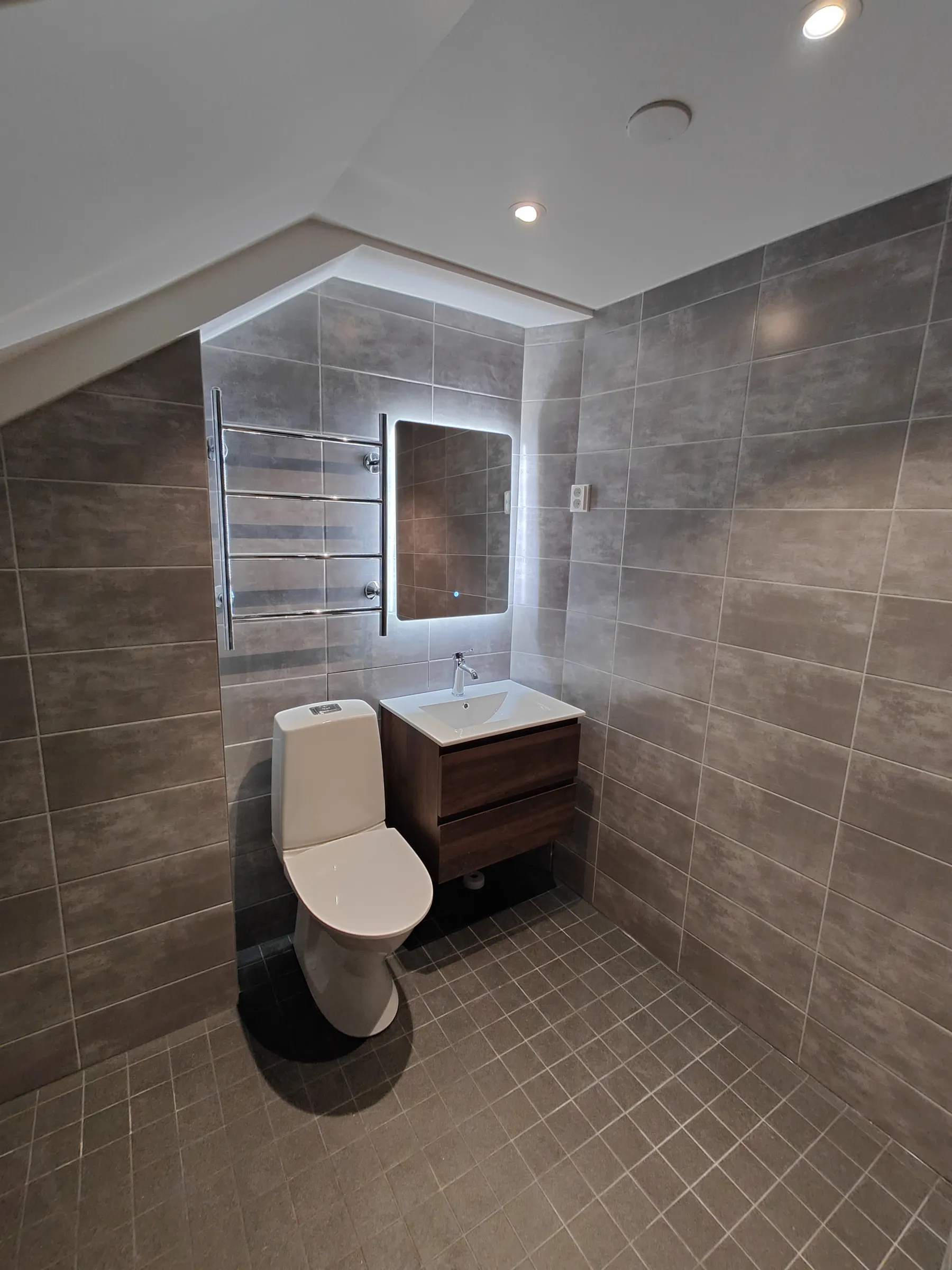 Moderni kylpyhuone harmailla laatoilla, valkoisella WC-istuimella ja puusävyisellä pesuallaslaatikostolla, peilissä valaistus.