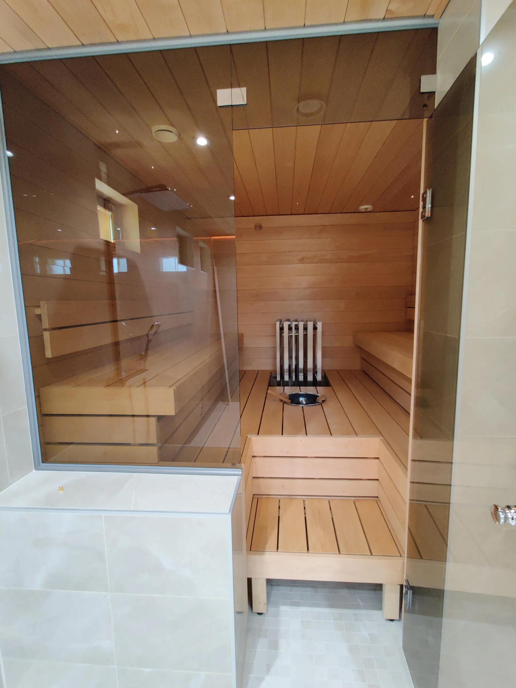 Sauna, jossa on puupanelointi, lasiovi ja puiset penkit, valaistus on kirkas ja lämmin.