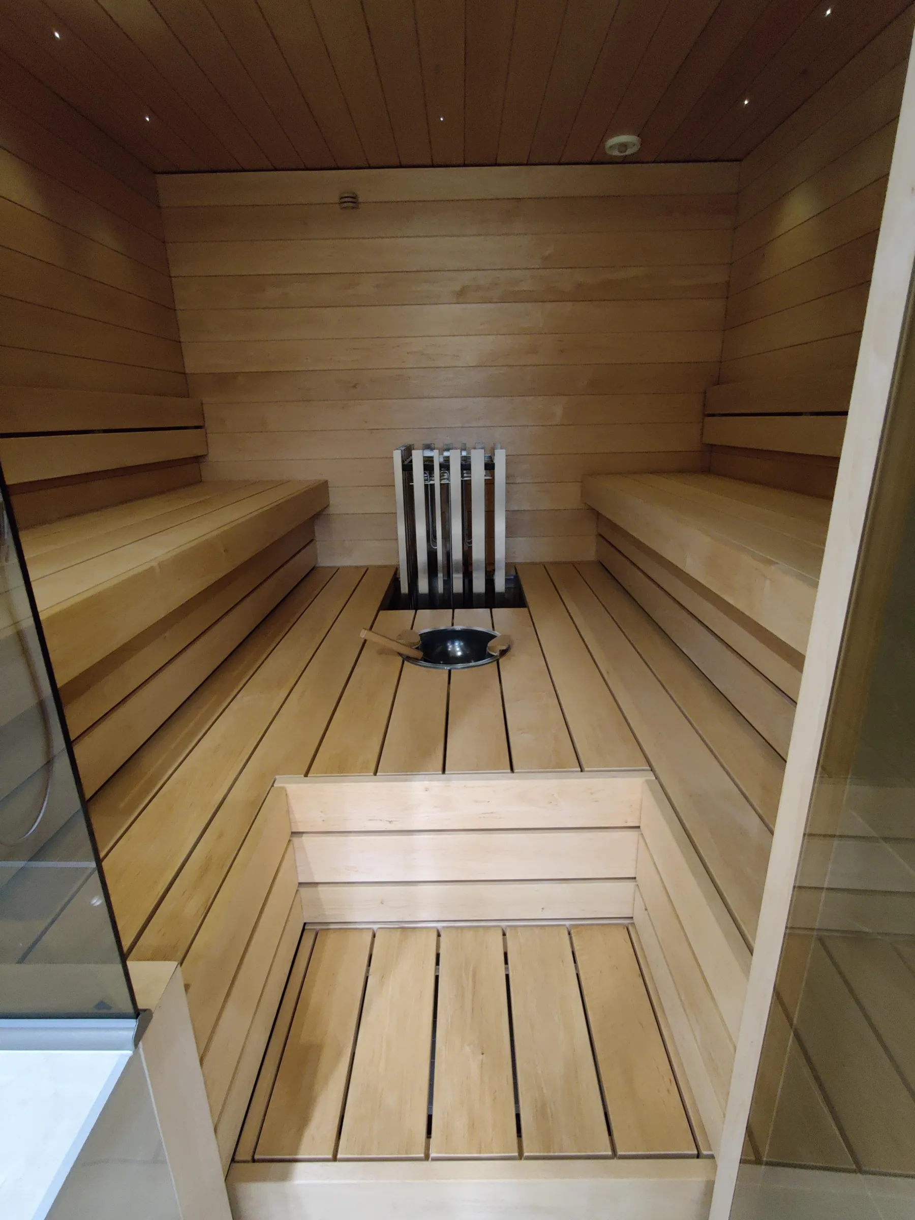 Puinen sauna, jossa on penkit, kiuas ja kauha. Valoisa ja lämmin tunnelma.