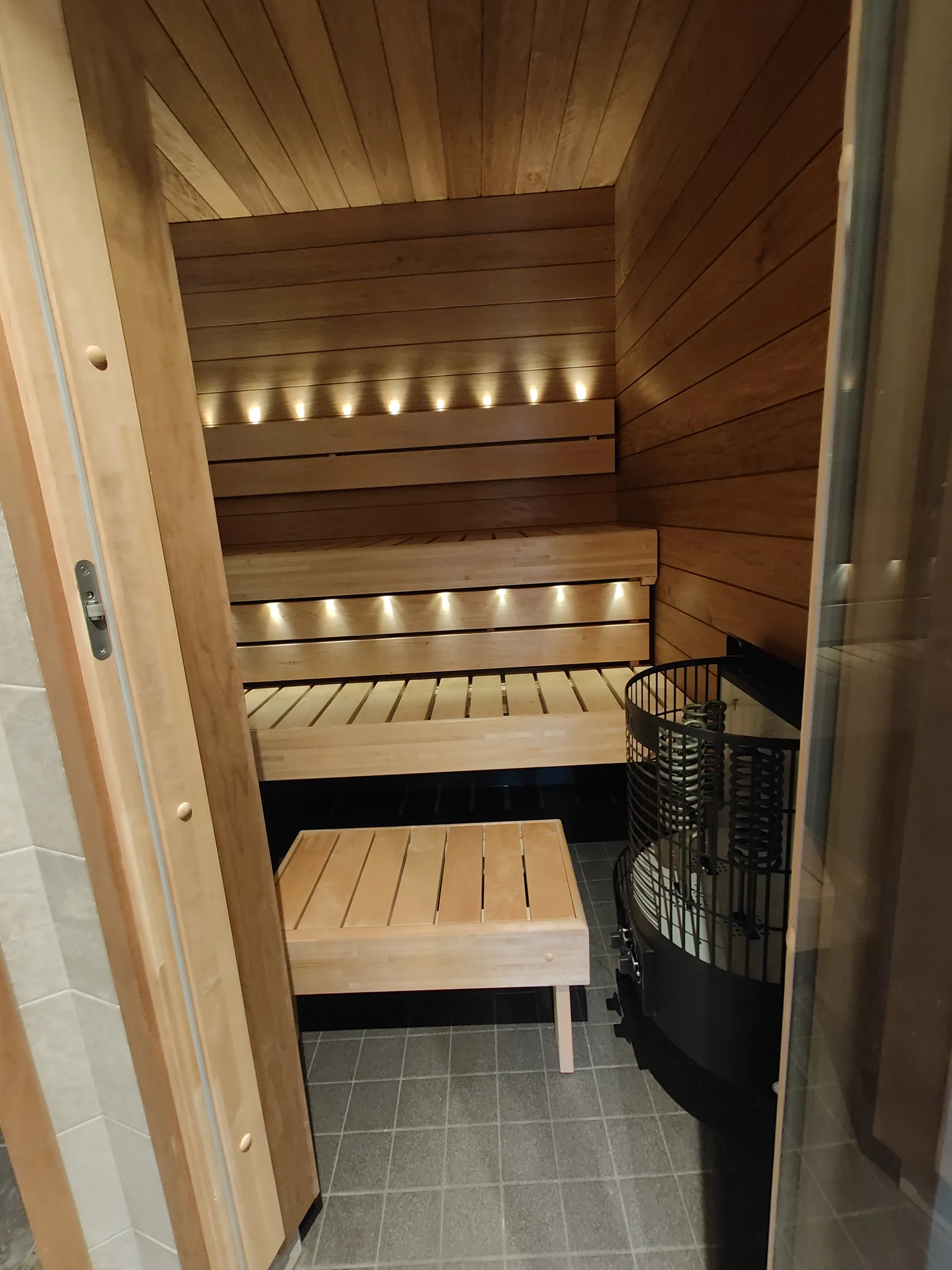 Sauna, jossa on puupanelointi, puiset penkit ja sähkökiuas, valaistu epäsuorasti.