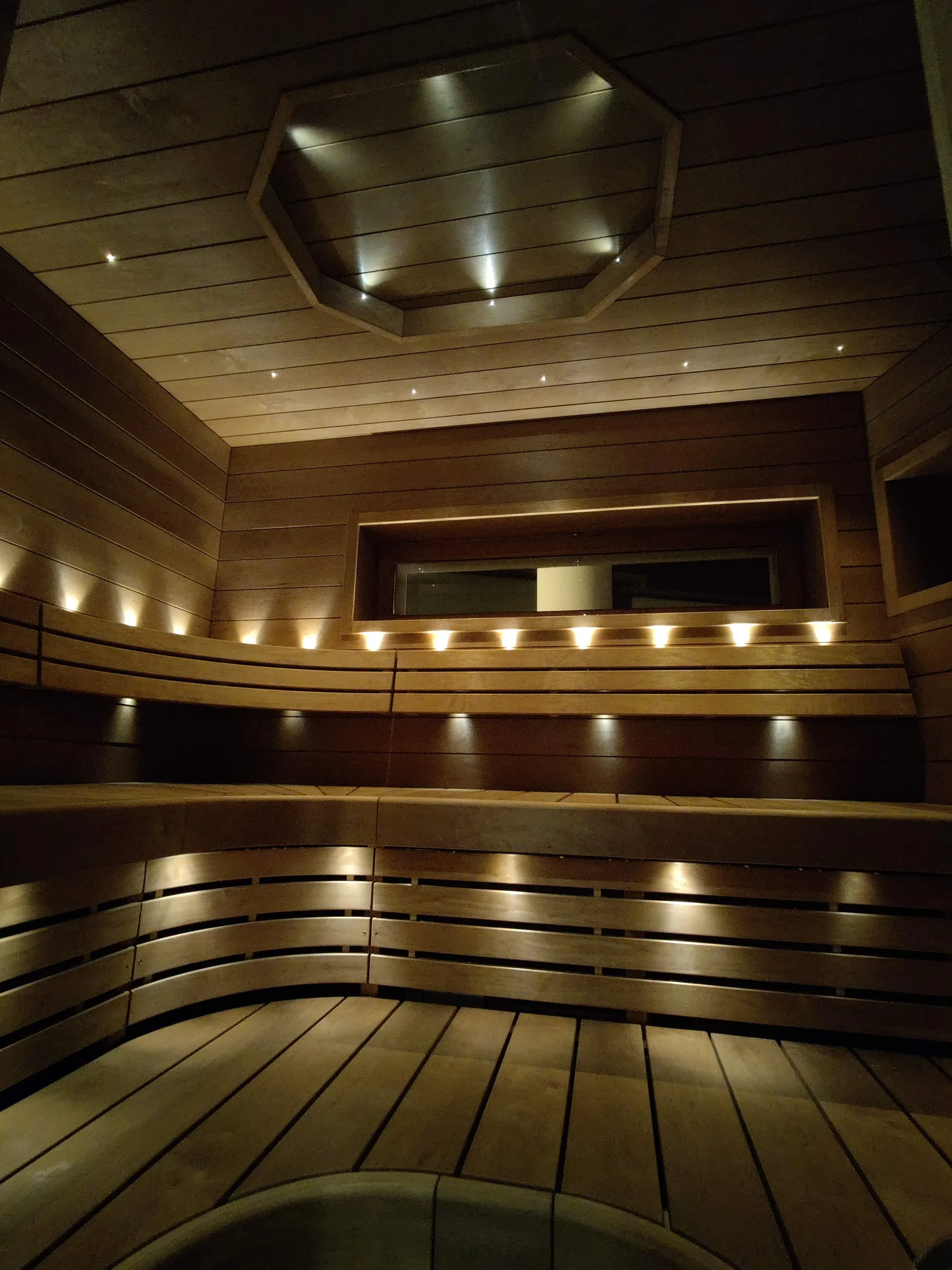 Sauna, jossa on puupanelointi, penkit ja epäsuora valaistus luovat lämpimän tunnelman.