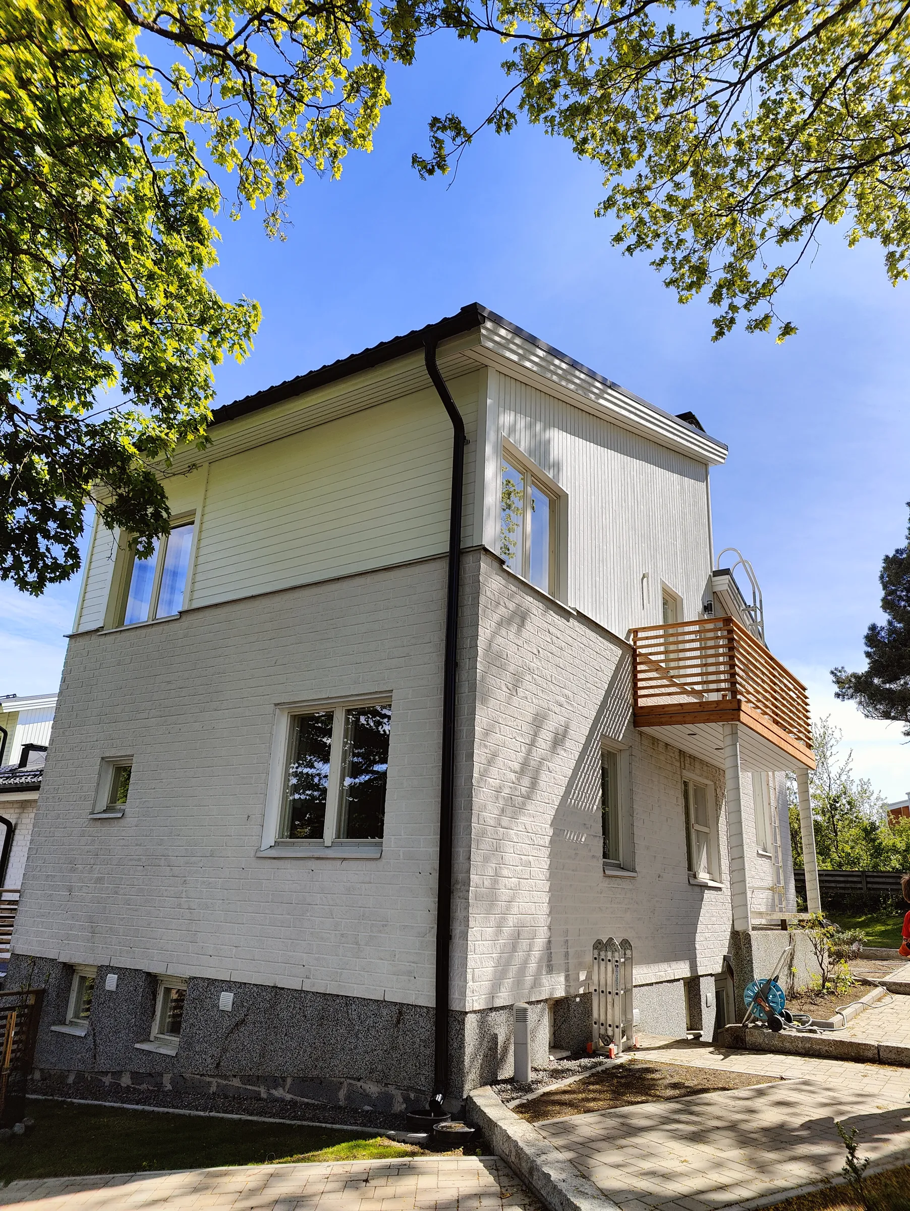 Kaksikerroksinen valkoinen talo, jossa on puinen parveke ja tiiliseinä, aurinkoisessa säässä.