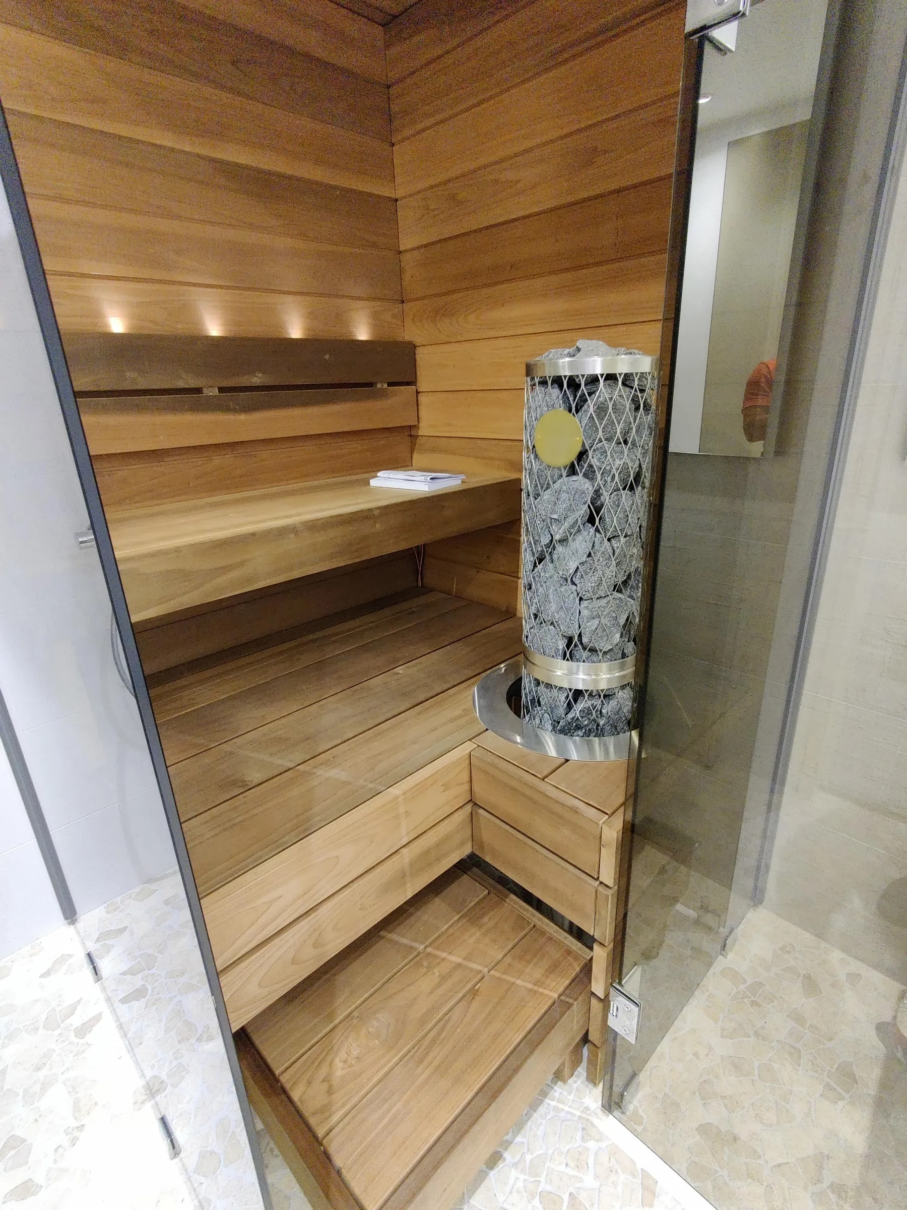 Puinen sauna, jossa on kivikiuas, penkit ja valaistus, sekä lasiovi ja marmorilattia.