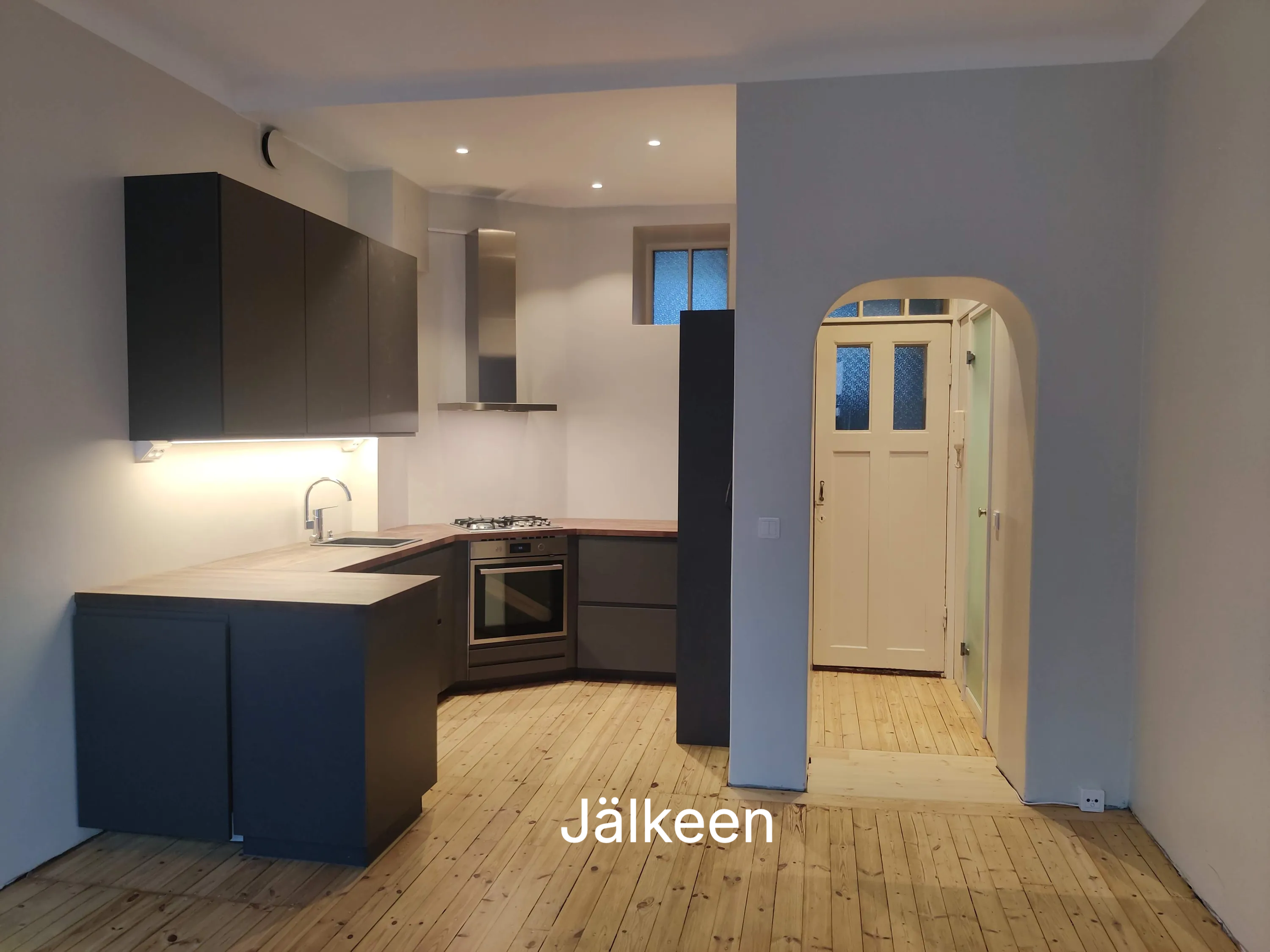 Moderni keittiö, jossa on tummanharmaat kaapit, puinen työtaso ja vaaleat puulattiat. Kaapistojen alla on valaistus ja huoneessa on pyöreä oviaukko.