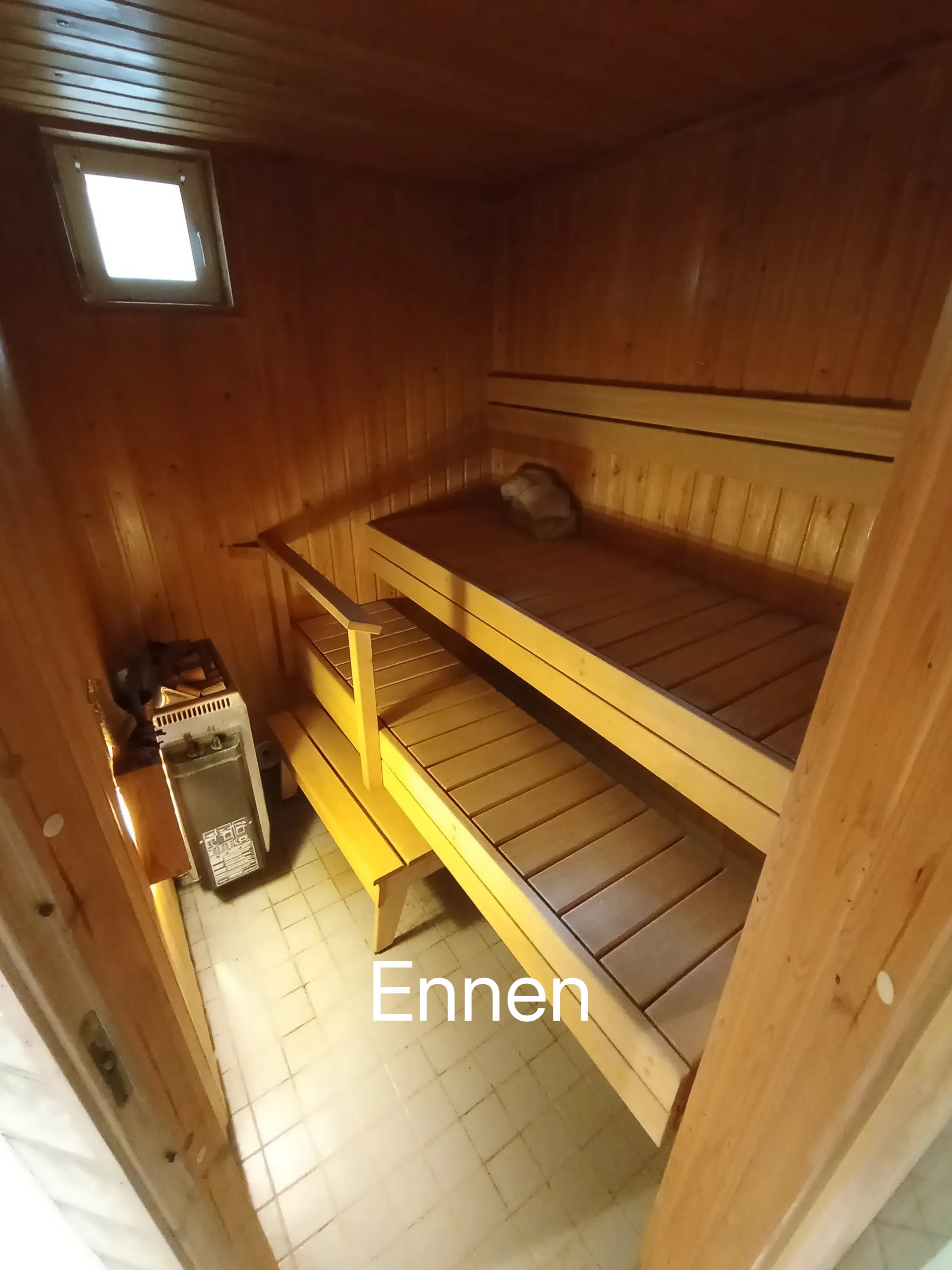 Puinen sauna, jossa on kaksi penkkiä, pieni ikkuna ja kiuas, valaistus on lämmin.