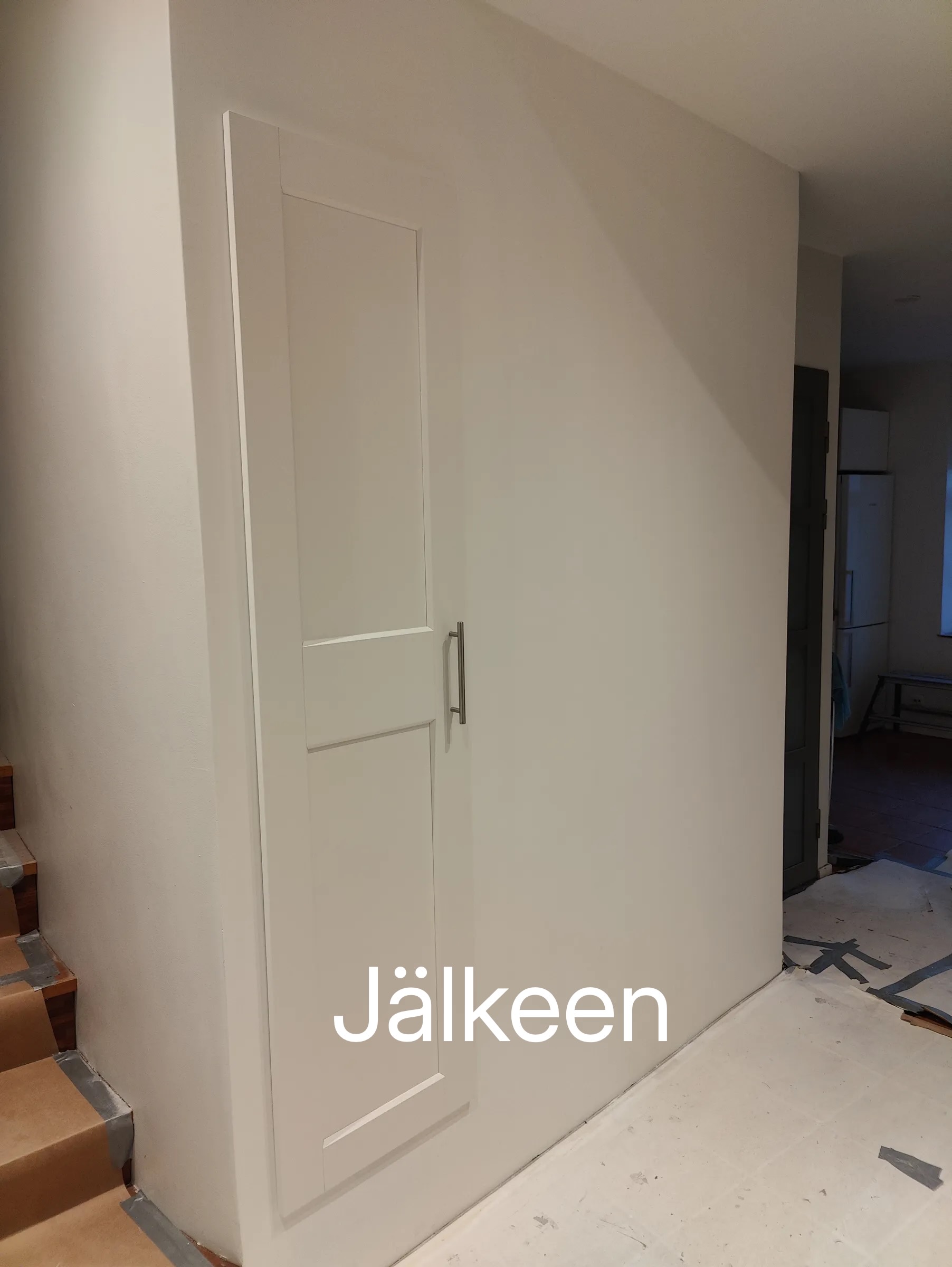 Valkoinen kaappi seinällä, metallinen kahva, portaat vasemmalla, valoisa huone.