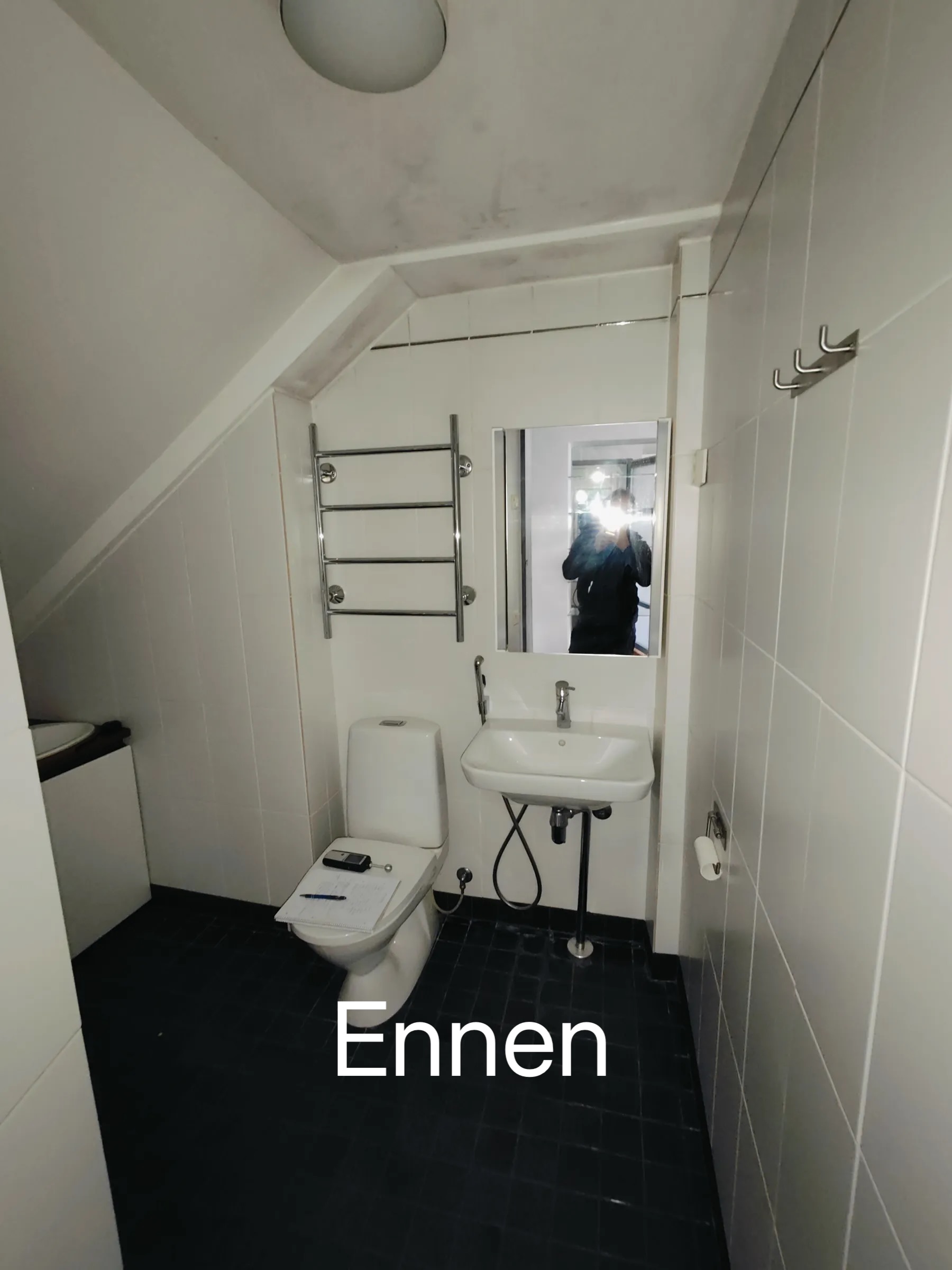 Kylpyhuoneessa on valkoinen wc, pesuallas ja pyyhekuivain, sekä tumma lattia ja valkoinen laatoitus.