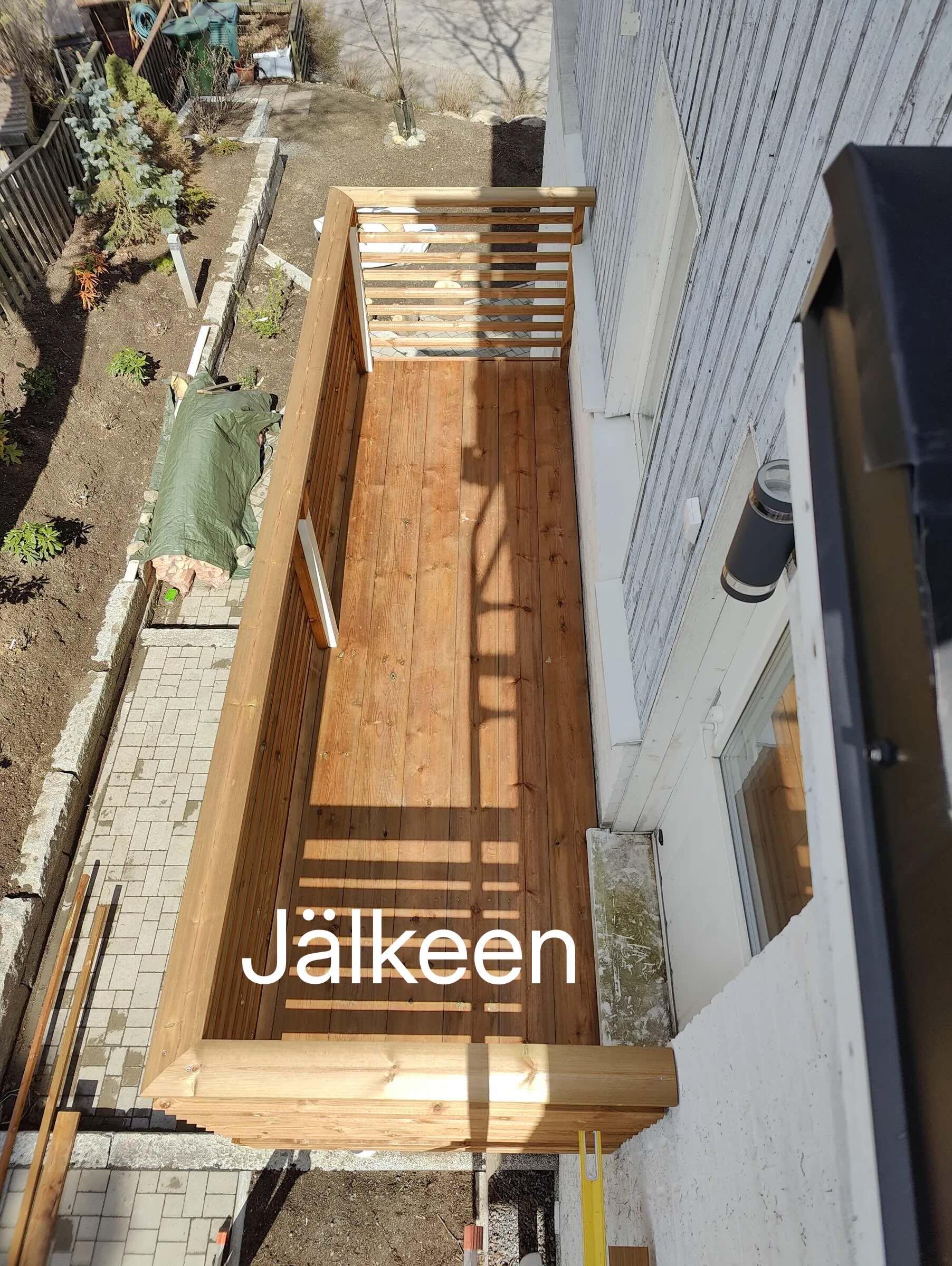Ylhäältä otettu valokuva puuterassista, jossa on puukaiteet ja puulattia. Terassi on rakennettu talon seinän viereen. Aurinko paistaa terassille ja luo varjoja.