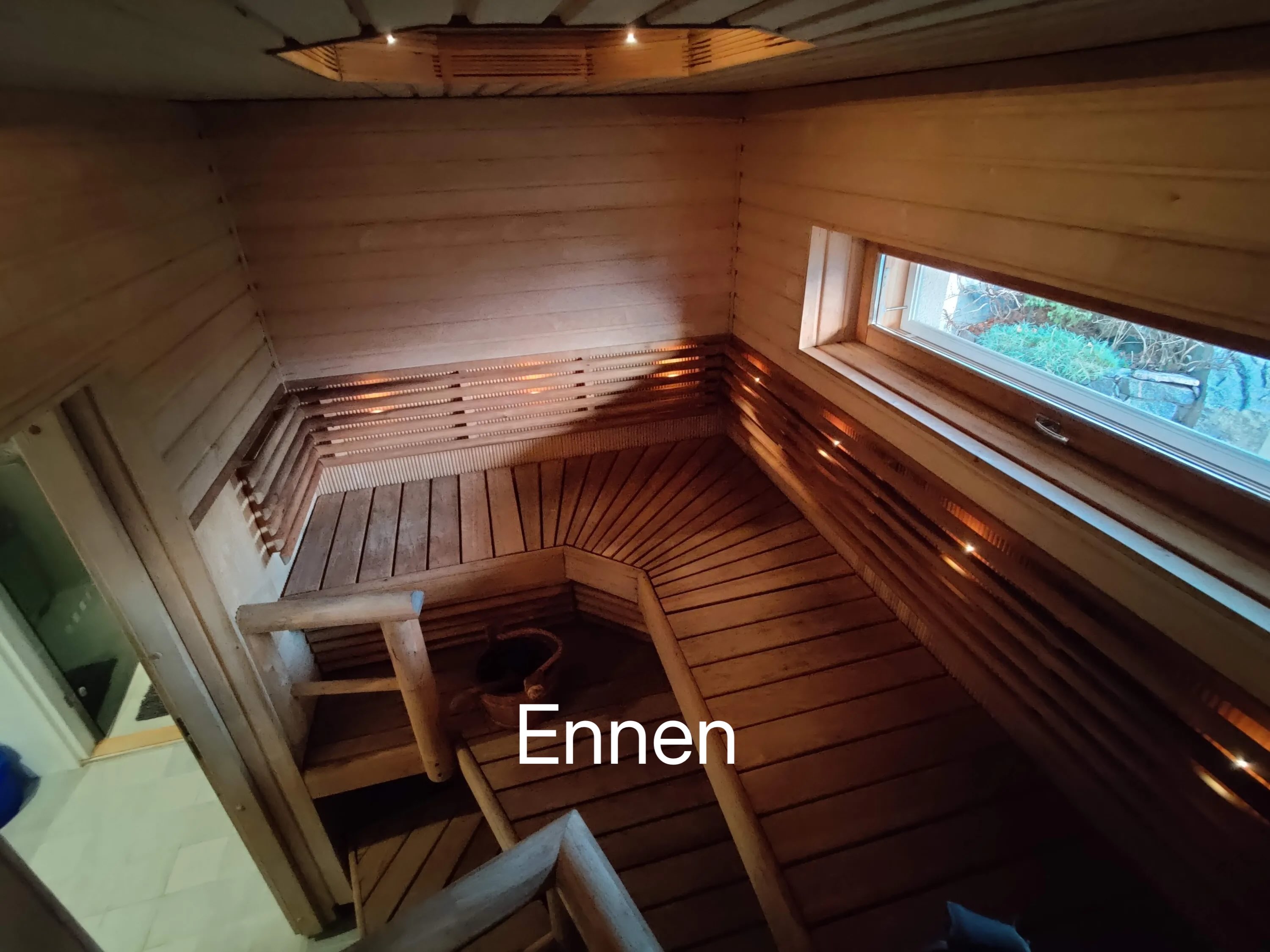 Puinen sauna, jossa on ikkuna, penkit ja kiulu. Valoisa ja lämmin tunnelma.