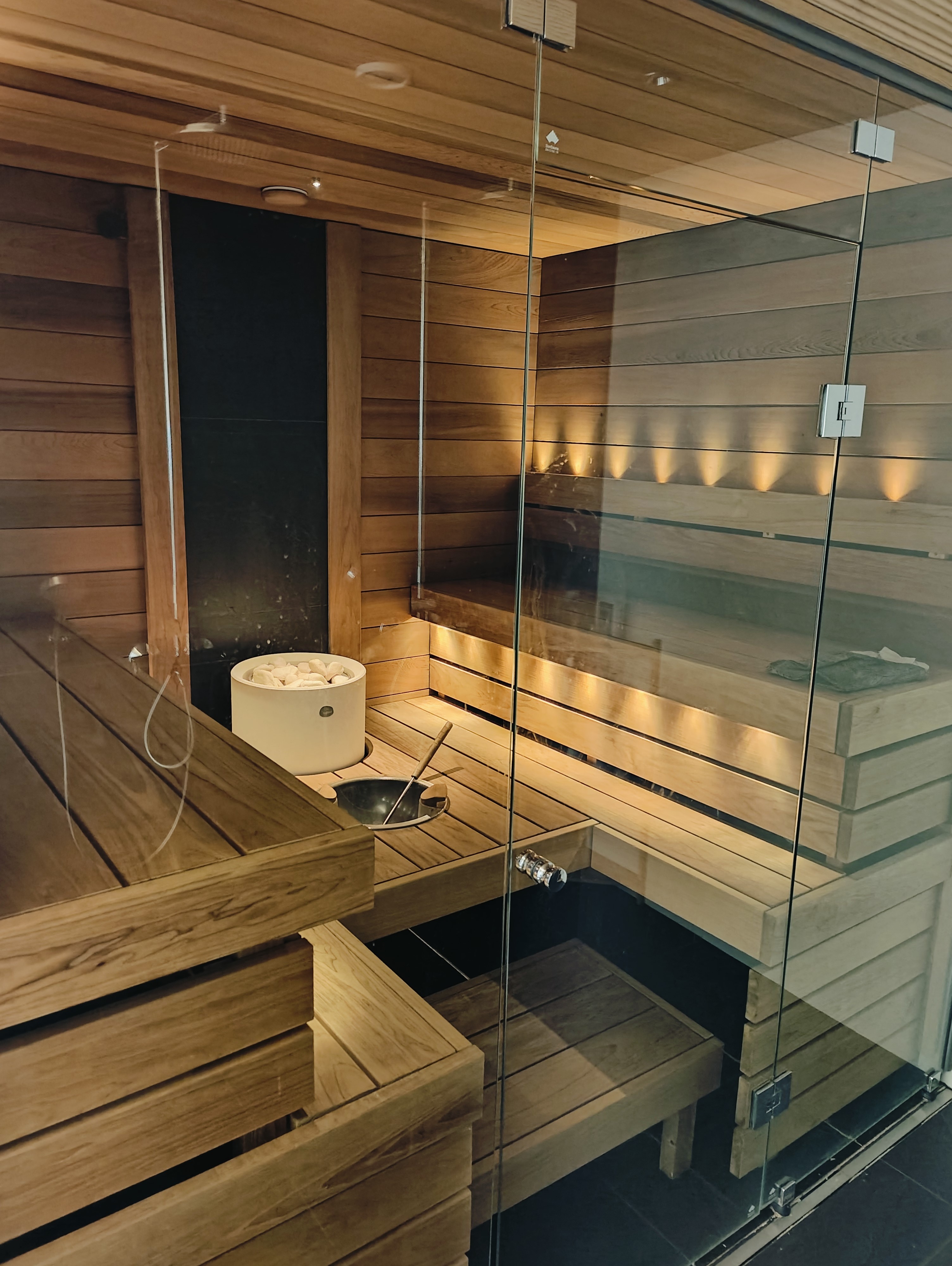 Sauna, jossa on puupaneelit, lasiovi ja valkoiset kivet lämmittimessä. Puu on vaaleanruskeaa ja valaistus on lämmin.