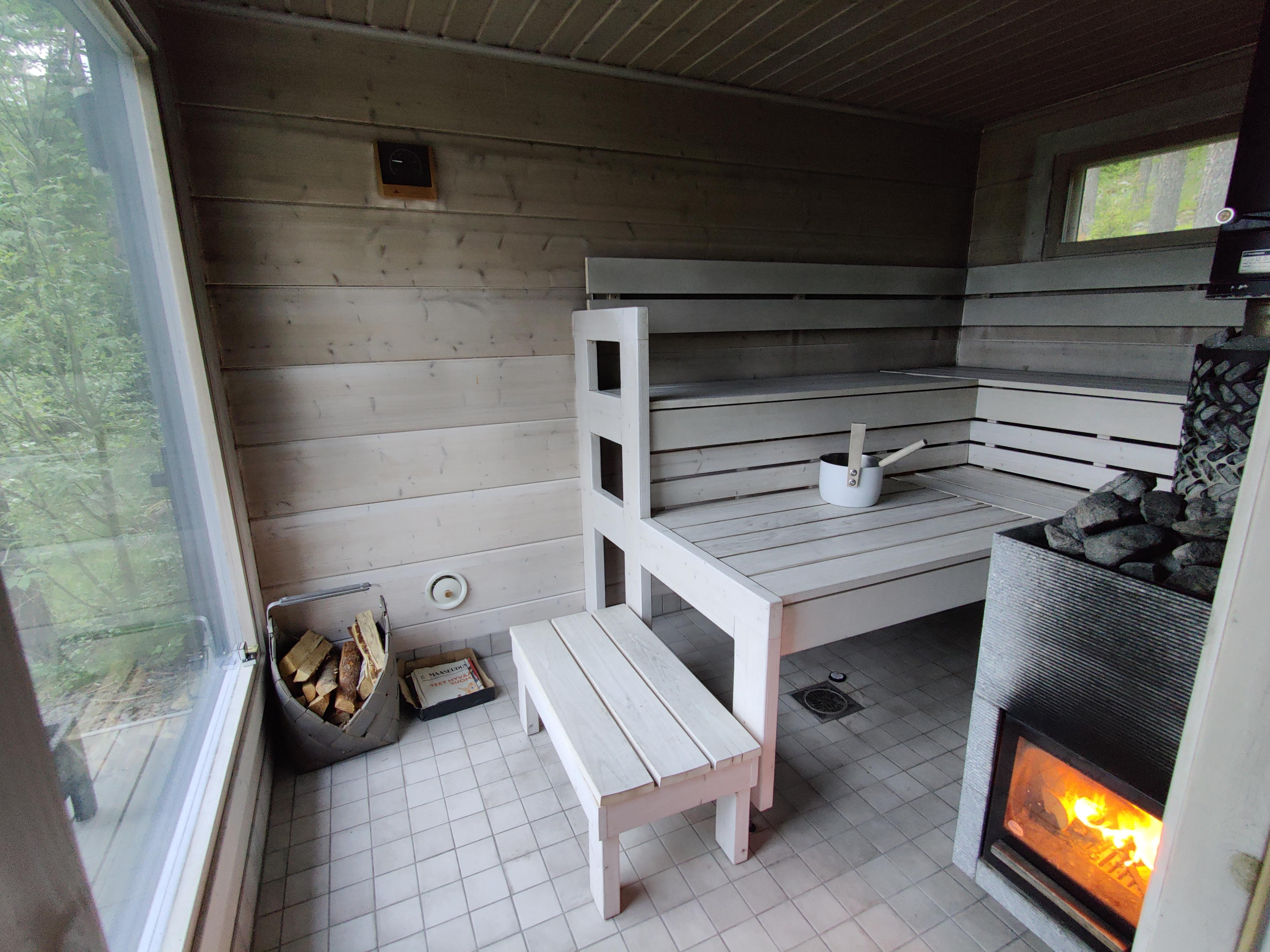 Sauna, jossa on puulämmitteinen kiuas, valkoiset lauteet, ikkuna ja puukori.