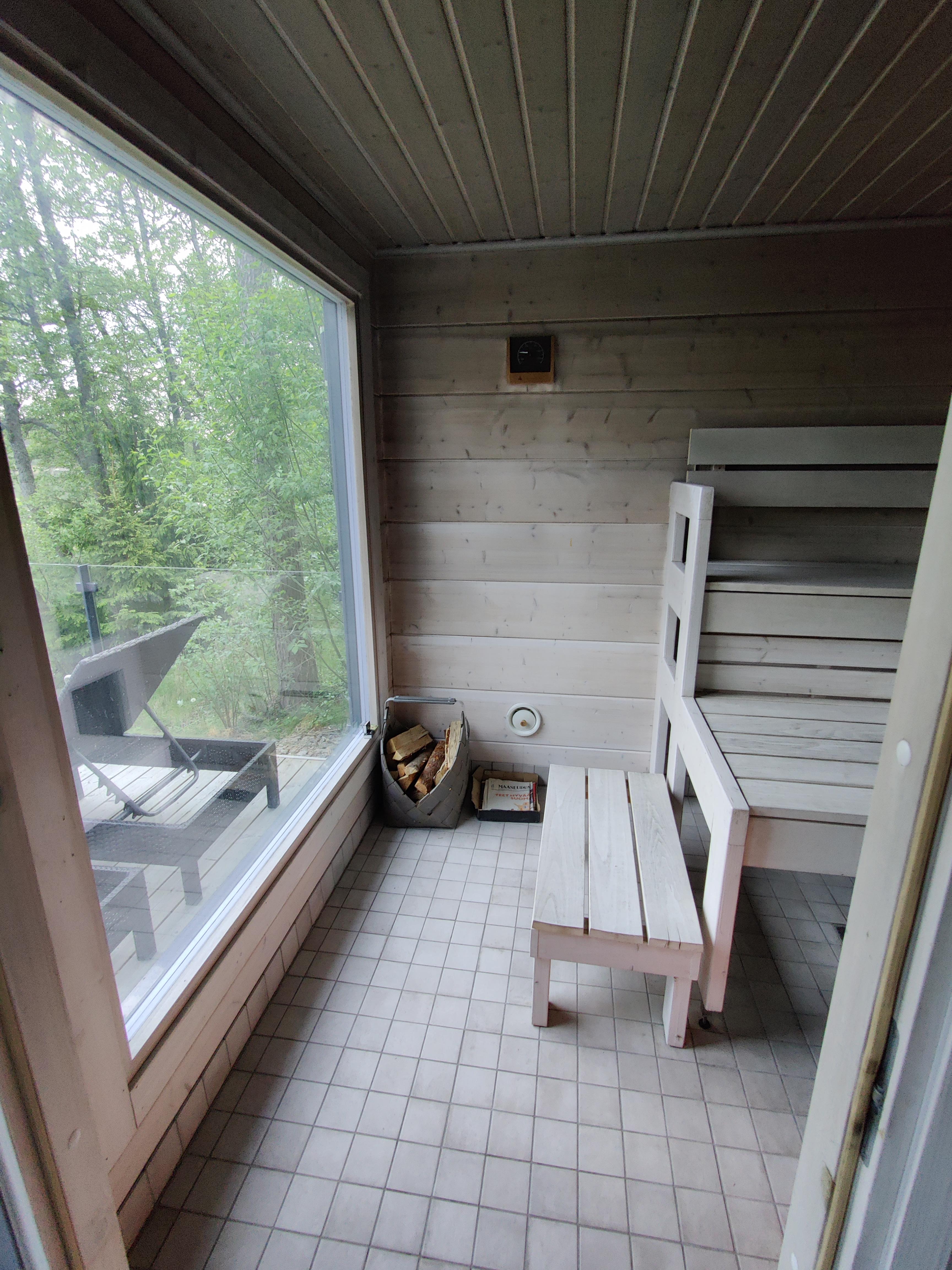 Sauna, jossa on puupanelointi, suuri ikkuna, puukori, penkki ja laattalattia. Valoisa ja rauhallinen tunnelma.