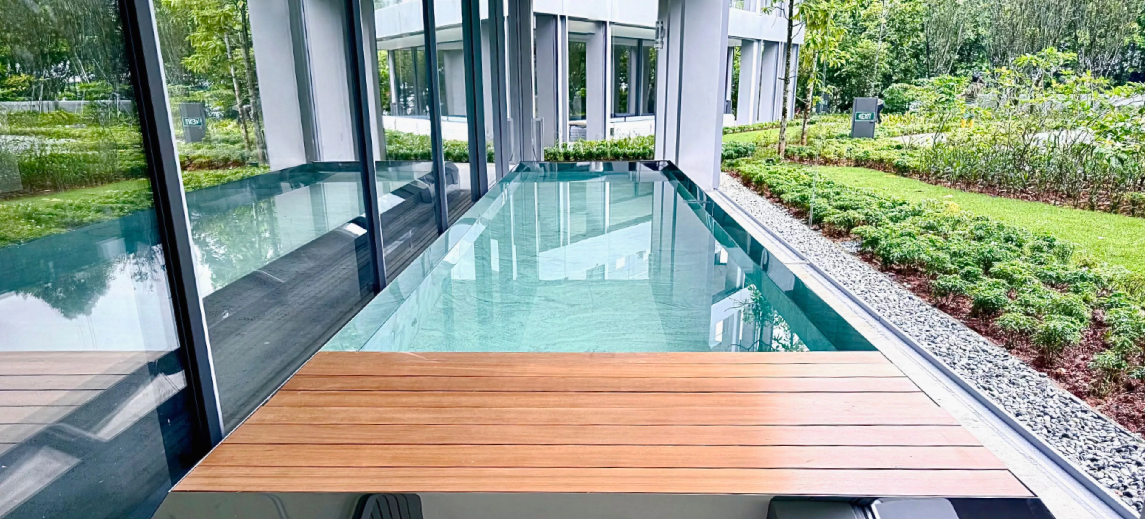 The Les Maisons lap pool in Singapore, with wooden planks on one end. Glass windows line the pool on one side, and green bushes on the other.