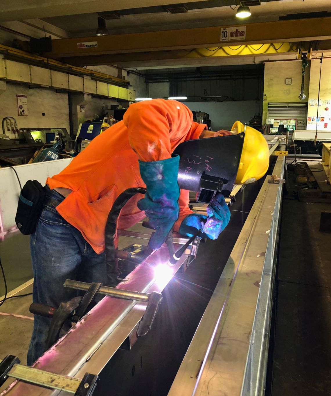 A worker wears protective face equipment, as bright sparks fly from a stainless steel piece he is welding. The background looks to be within a construction factory.