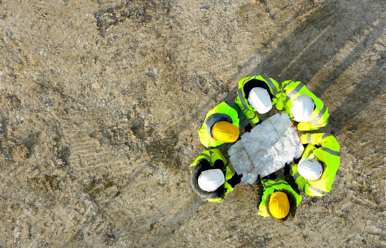 A group of construction workers in bright yellow safety work gear are huddled together, peering over a construction plan.