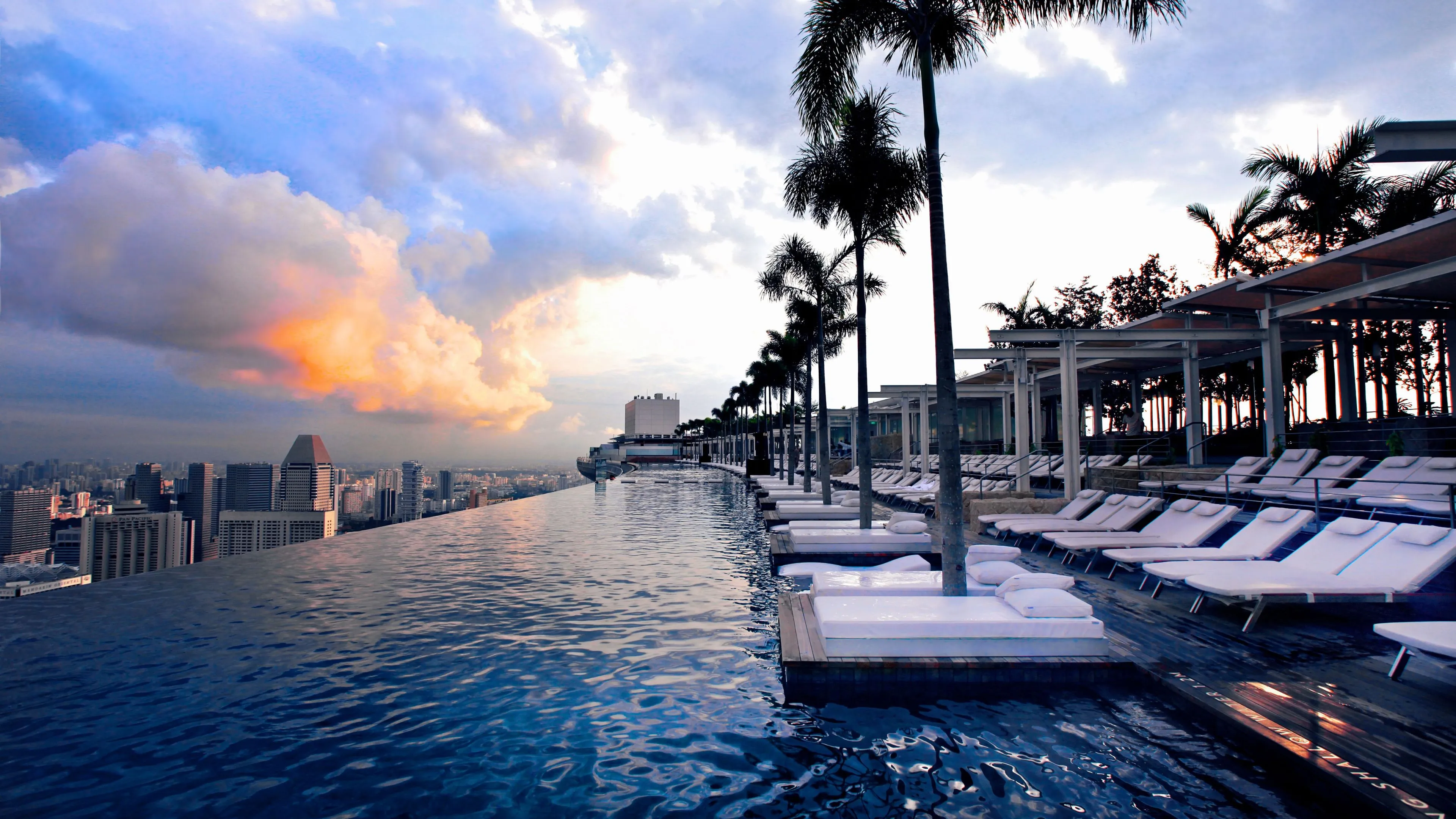 The Marina Bay Sands SkyPark infinity pool glistens under a serene sky at dusk. It faces outwards towards the central business district of the Singapore skyline, with empty deck chairs on the opposite end.