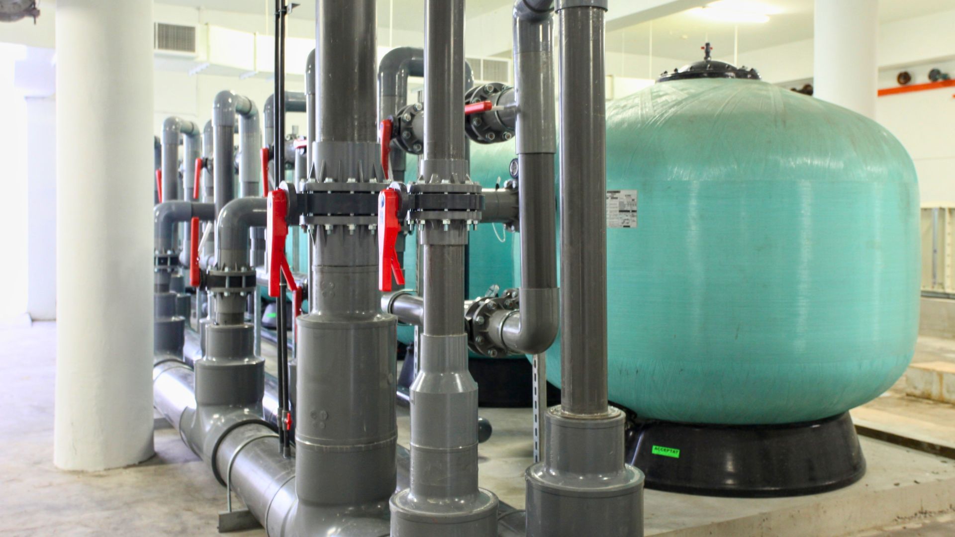 Close-up of the green high-rate fibreglass pool filters, grey UPVC piping, and bright red valves in the swimming pool pump room at Anglo-Chinese School Independent (ACSI), Singapore.