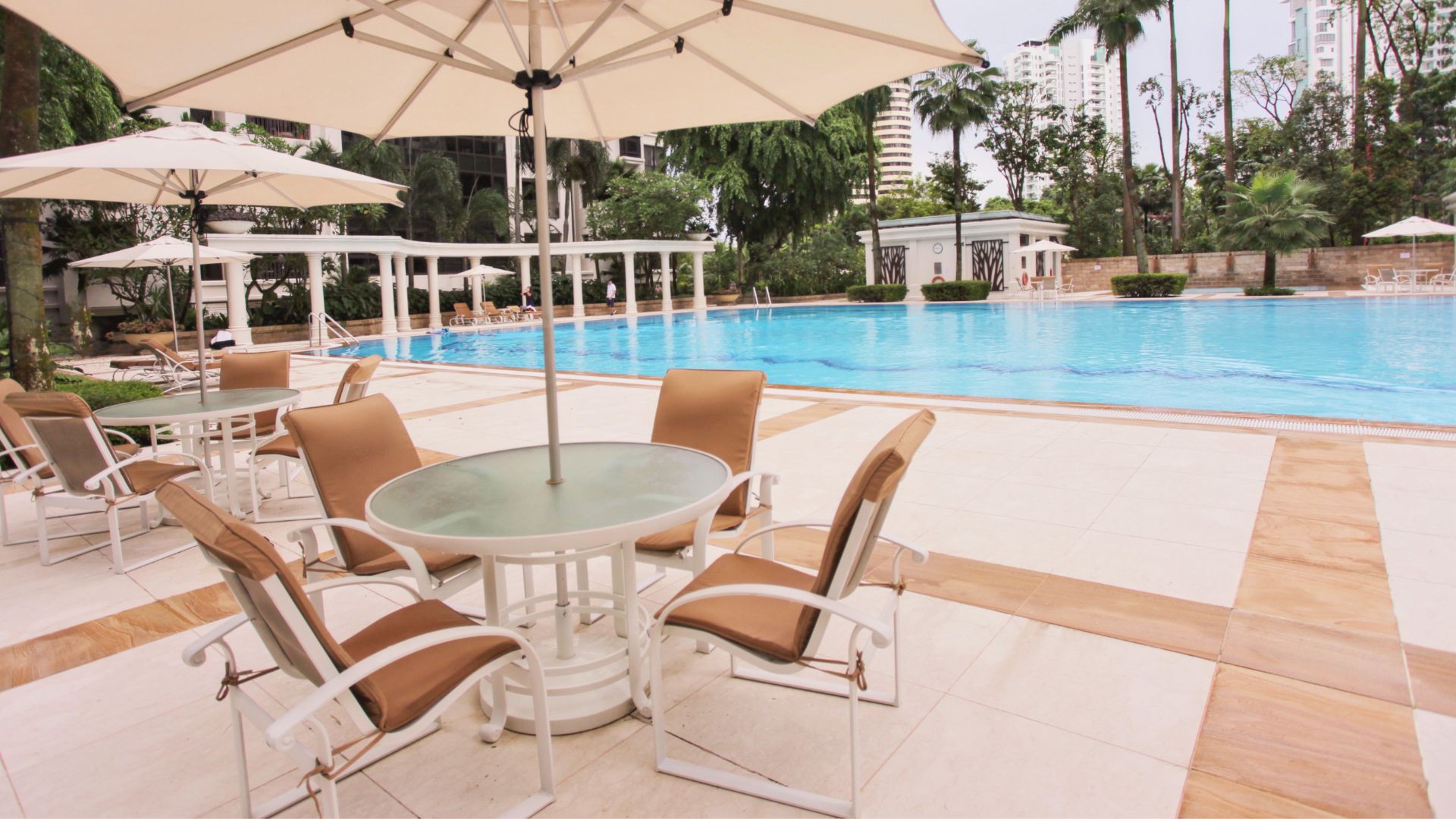 The pool deck of Ardmore Park condominium in Singapore. Tables and chairs are scattered in clusters, with ample space between them. A large white umbrella provides shade above each table, and the chairs are lined with water-absorbent padding in brown.