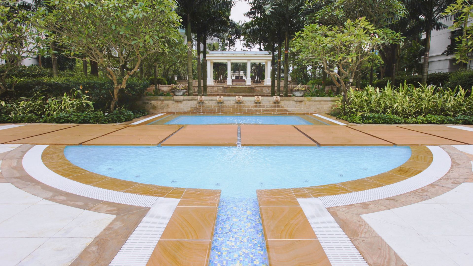 The stainless steel jacuzzi pool at Ardmore Park, surrounded by greenery. A granite walkway divides it from water features in the background. Blue tiles sparkle in a narrow waterway in the foreground, before opening into the wide, circular jacuzzi area.
