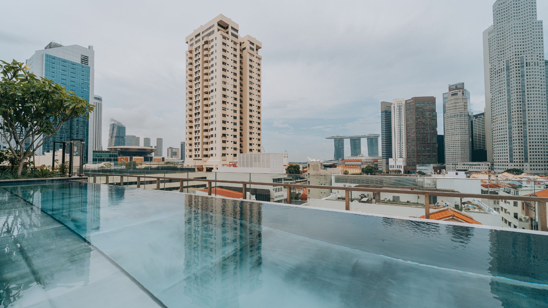 Eye-level view of the swimming pool at 21 Carpenter hotel, with a panoramic view of Singapore city. Tall, modern buildings intersperse with short, traditional complexes.