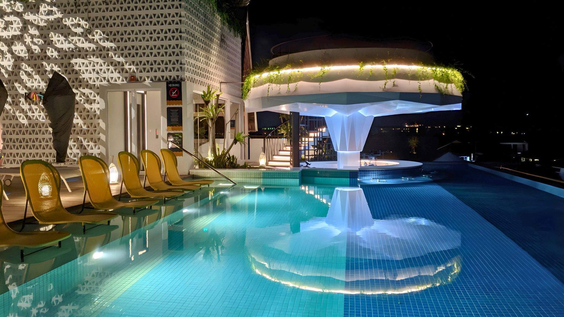 The stainless steel rooftop pool at Aloft Bali Seminyak, Marriott hotel, brightly lit up at night. Empty rattan deck chairs line up in a row, with a submerged bar at the end.