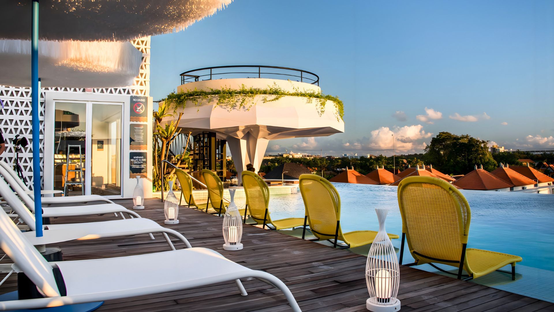 The rooftop pool and pool deck at Aloft Bali Seminyak — Marriott, with a submerged bar. There are two rows of deck chairs aligned to face the pool. The first row of rattan chairs sit right above the water, and the white chairs behind them are adjustable.