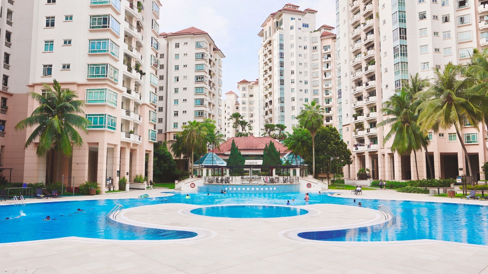Panoramic view of Costa Rhu condominium in Singapore. The large, resort-style swimming pool stands out with a curvaceous concrete walkway that leads to a central, circular area. The residential blocks in the background overlook the pool.