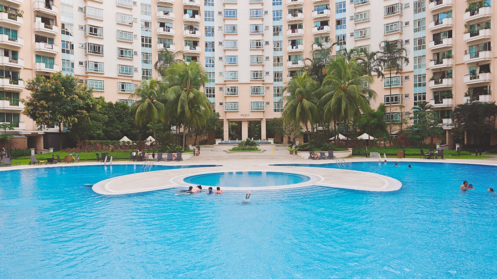 Panoramic view of the swimming pool at Costa Rhu condominium, facing Ancilla block. The landscape is lush with greenery, with a spacious pool deck. A wide, concrete walkway leads into the pool in a circular shape, with pool steps at the other end.