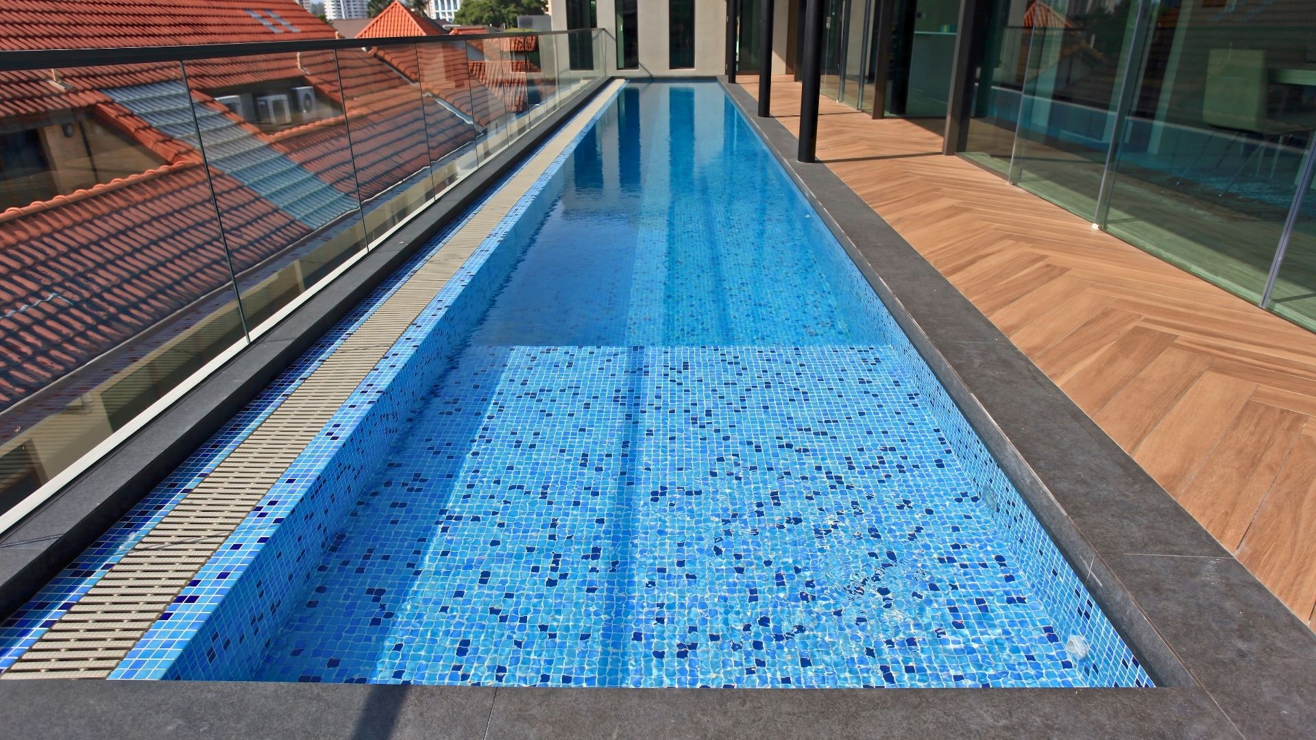 Eye-level view of the rooftop swimming pool at a private landed property in Singapore. Blue and white mosaic tiles line the pool floors and sides, and the narrow corridor with herringbone-patterned wood flooring beside it.