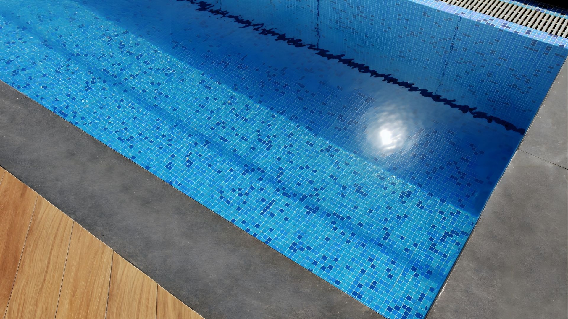 Close-up of the stainless steel pool at a private landed home in Singapore. Wooden flooring gives way to elegant, dark grey slabs which enclose the pool. Blue and white mosaic tiles line the inner side of the swimming pool.