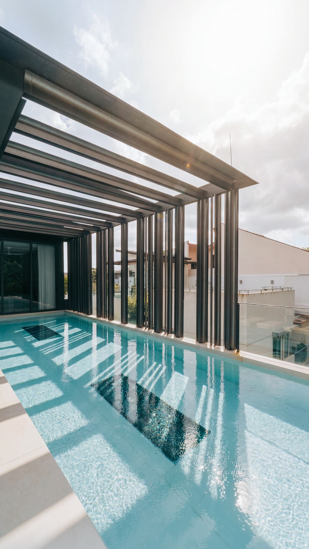 Portrait view of a stainless steel rooftop pool with underwater windows, facing towards the interior of the house. Beautiful overhead retractable shades, with sunlight streaming through the open gaps.
