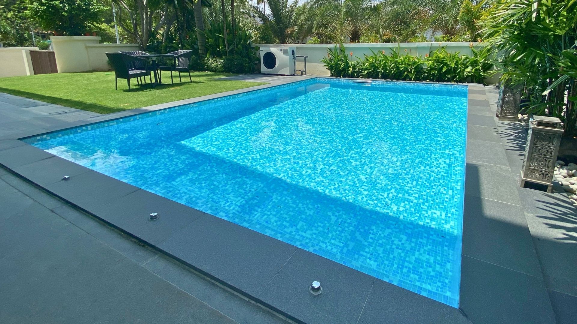 A stainless steel in-ground pool blends seamlessly into the garden landscape of a private landed home in Singapore. A well-trimmed lawn with a table and chairs is located right next to the pool.