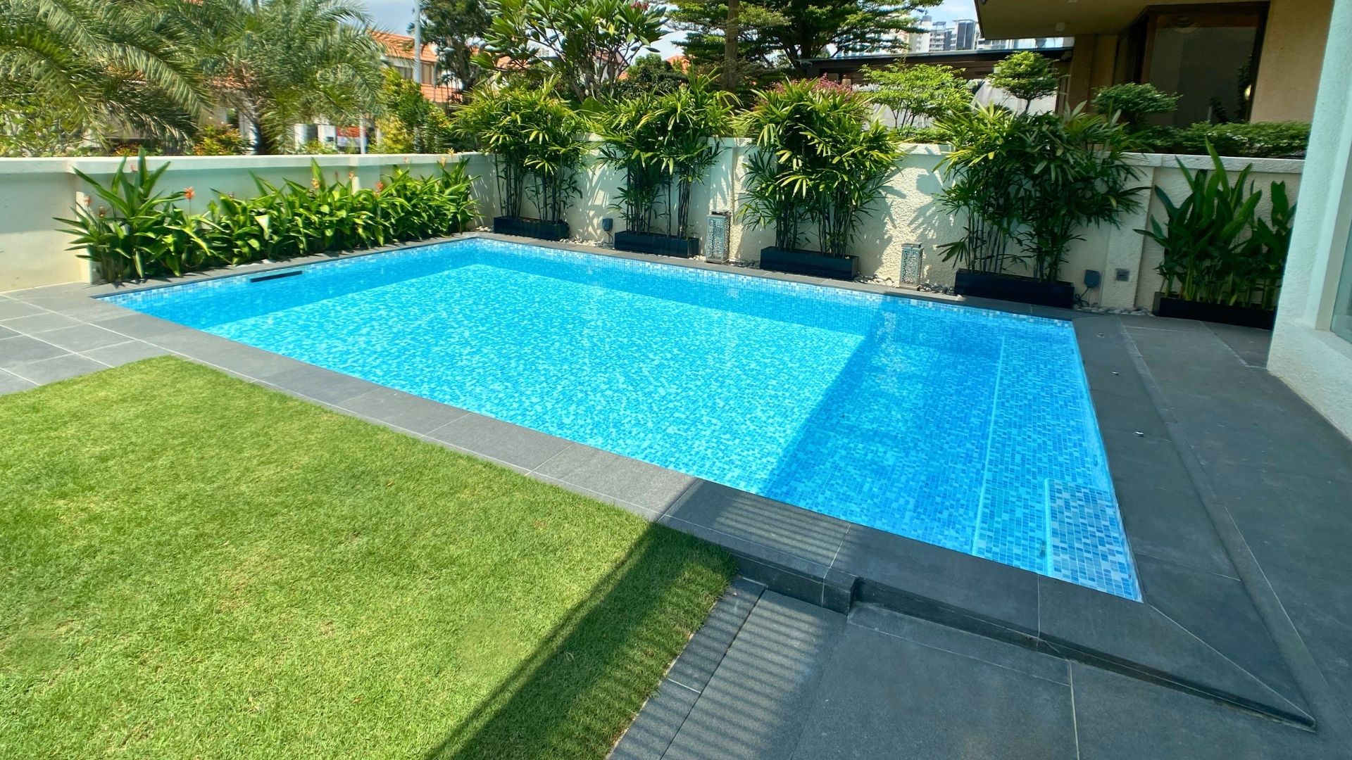 An in-ground swimming pool built within the garden of a private landed home in Singapore. The area around it is surrounded by dark-grey slabs of stone, with yet more space in the garden for plants and flowers.