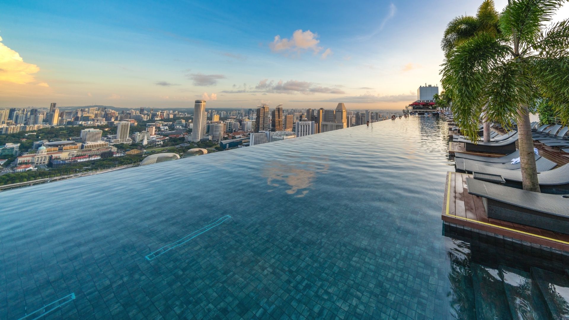 The iconic SkyPark Infinity Pool at Marina Bay Sands, with a sleek edge that seems to vanish. Individual deck chairs have been built into wooden sections, and face out directly for a full view of the Singapore skyline.