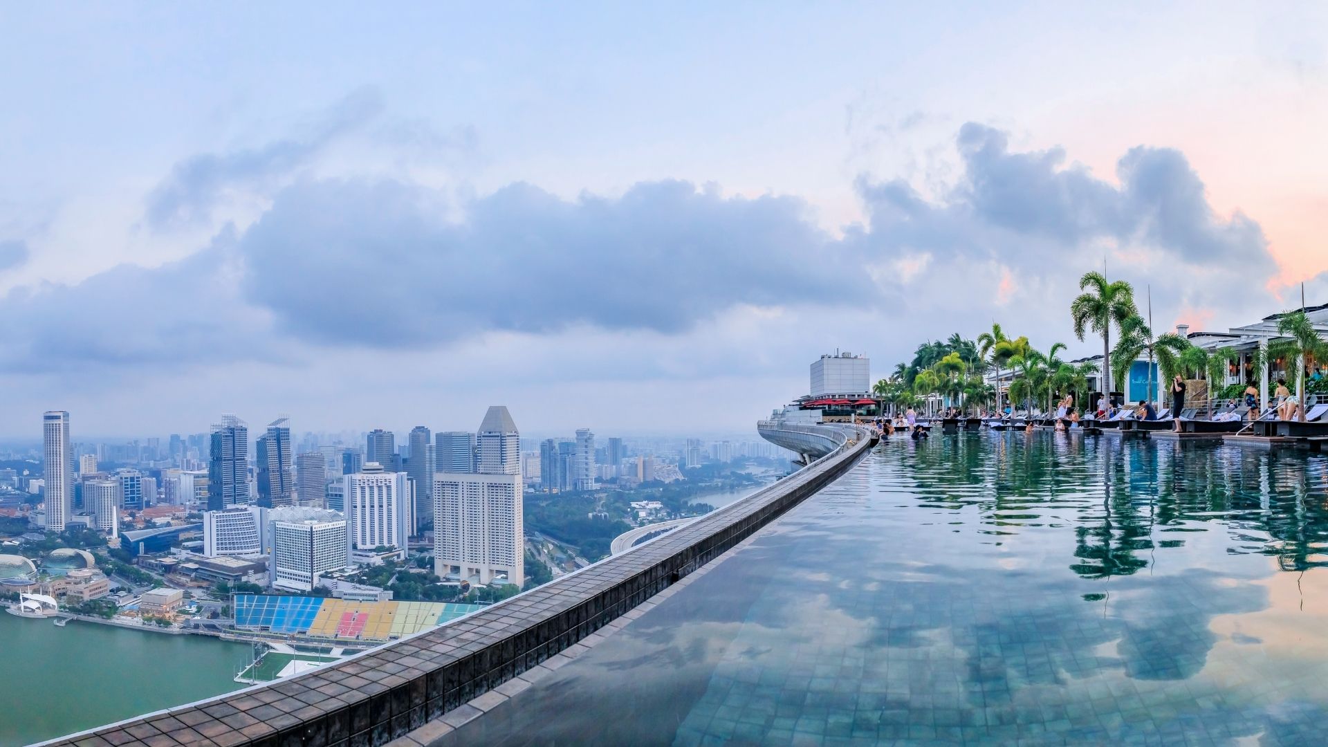 Eye-level view of the SkyPark Infinity Pool at MBS (Marina Bay Sands). Water overflows into the narrow pool drain at the edge, and Singapore city can be seen from a top-down view. People are relaxing in the deck chairs or in the swimming pool.