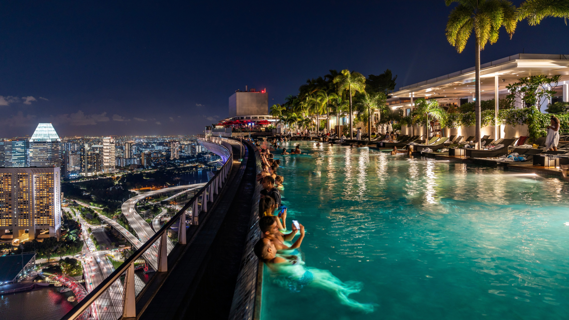 The Marina Bay Sands SkyPark Infinity Pool lit up at night, from underwater lights and warm ambient lighting at the pool deck. People in the pool lean against the infinity edge, with a backdrop of Singapore city twinking behind them.