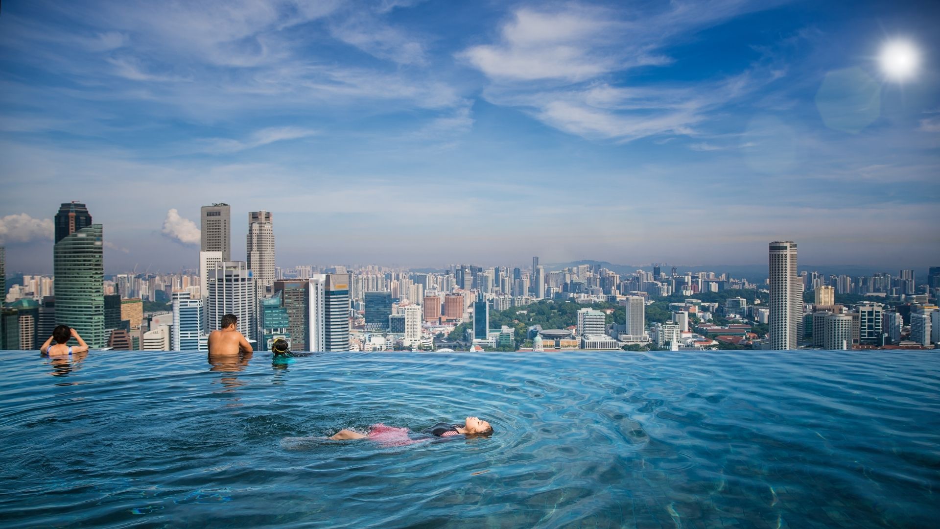 Sunlight reflects against the Marina Bay Sands SkyPark Infinity Pool. A girl floats in the middle of the pool on her back with her eyes closed, looking relaxed. A family leans against the infinity edge to take in the panoramic view of Singapore city.
