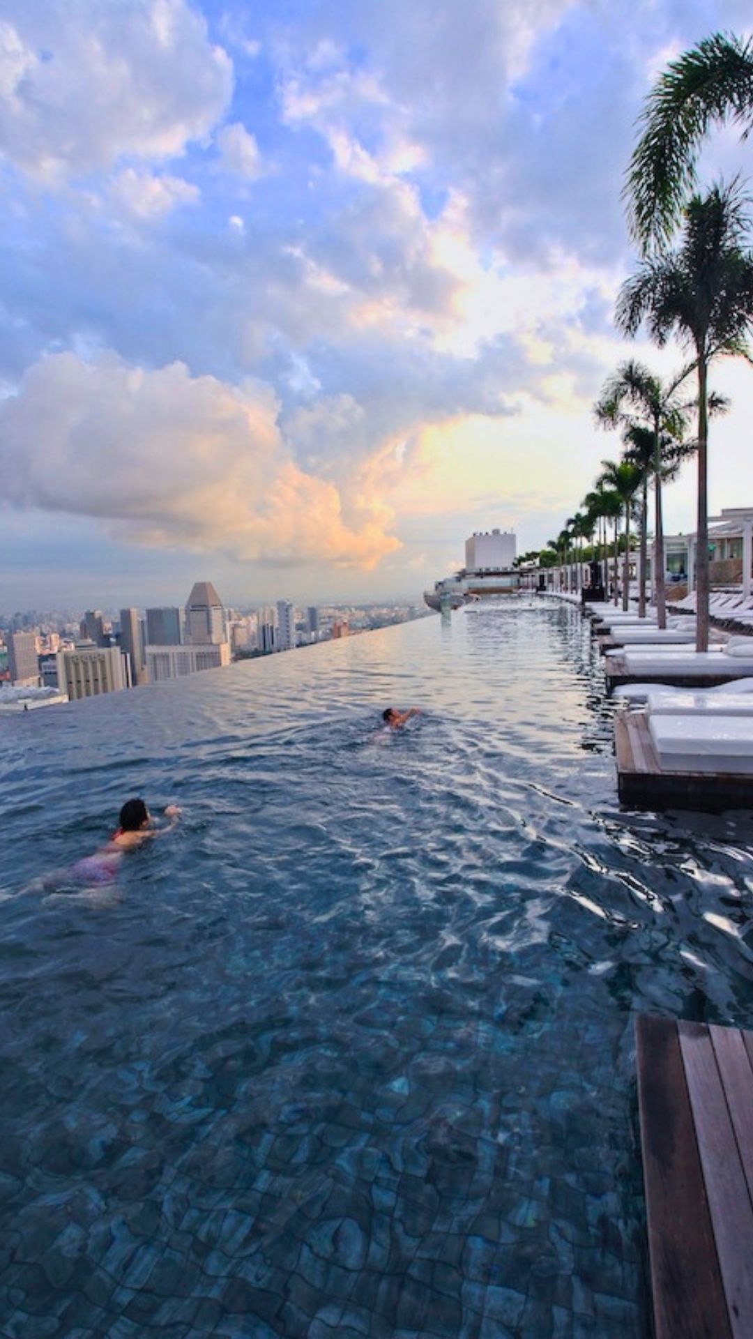 Swimmers enjoy a swim within the 150-metre long SkyPark Infinity Pool at Marina Bay Sands (MBS) in Singapore. The deck chairs to the right are empty as it appears to be dusk.