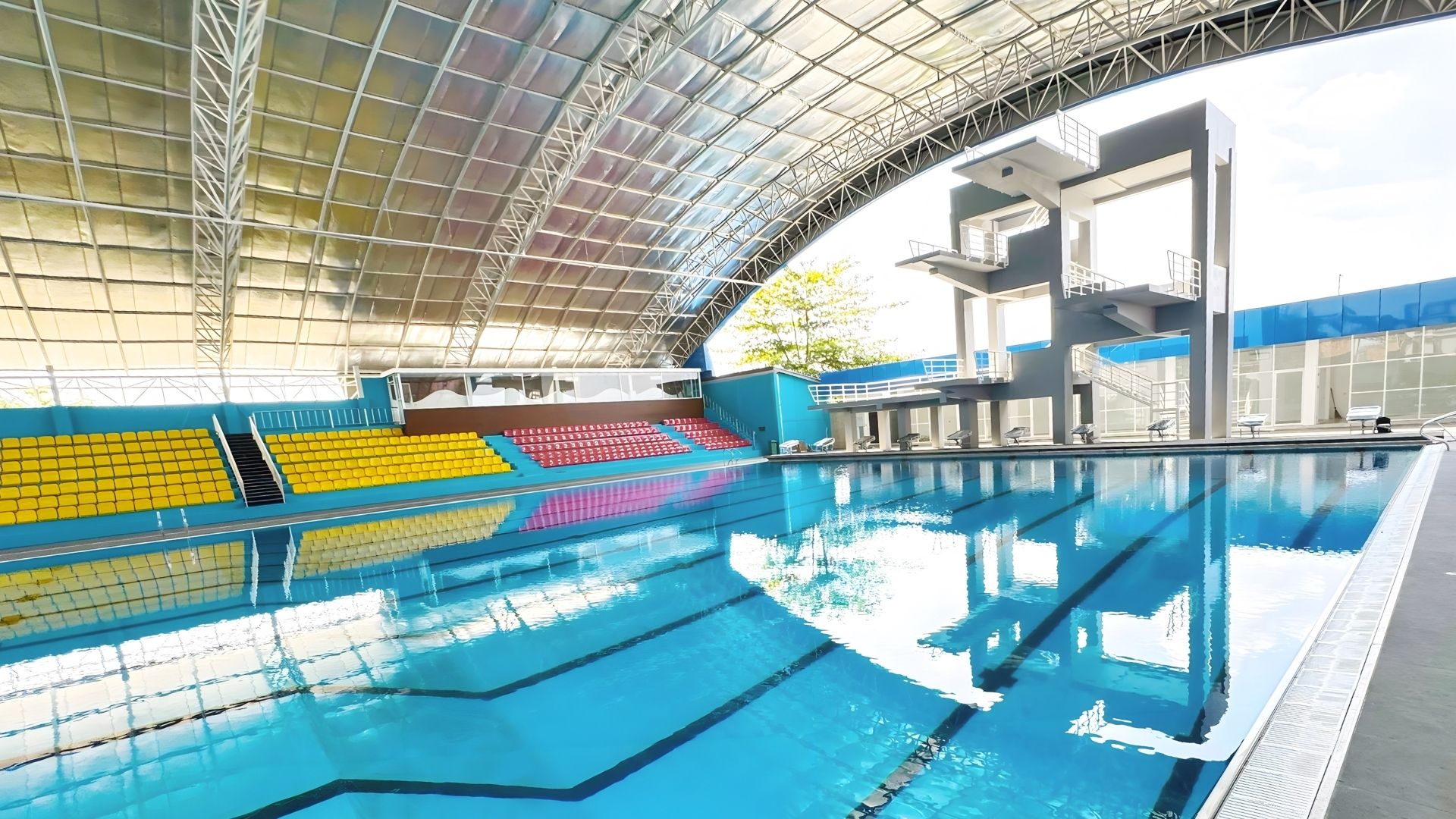 Panoramic view of the Selayang Aquatic Centre in Medan, which is enclosed indoors with metal structures. One side of the pool comprises starting blocks and diving boards. The competition pool is sloped in the middle from the shallow to deep end.