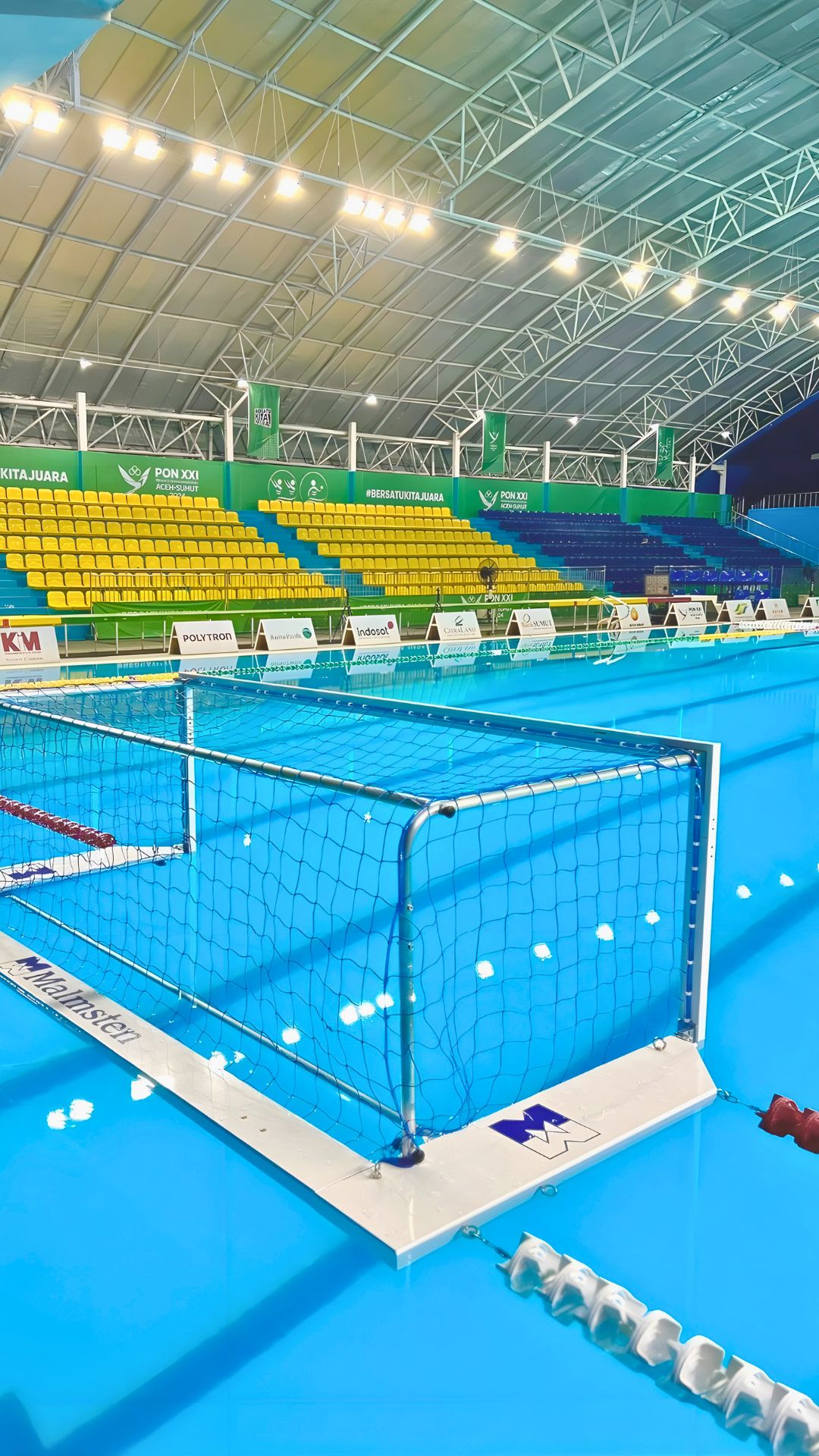 Close-up of the water polo net at the Selayang Aquatic Centre, fixed to colourful lane ropes. Spectator sections are divided in colour sections in the background. The logo of sponsors are printed on big white cards below them.