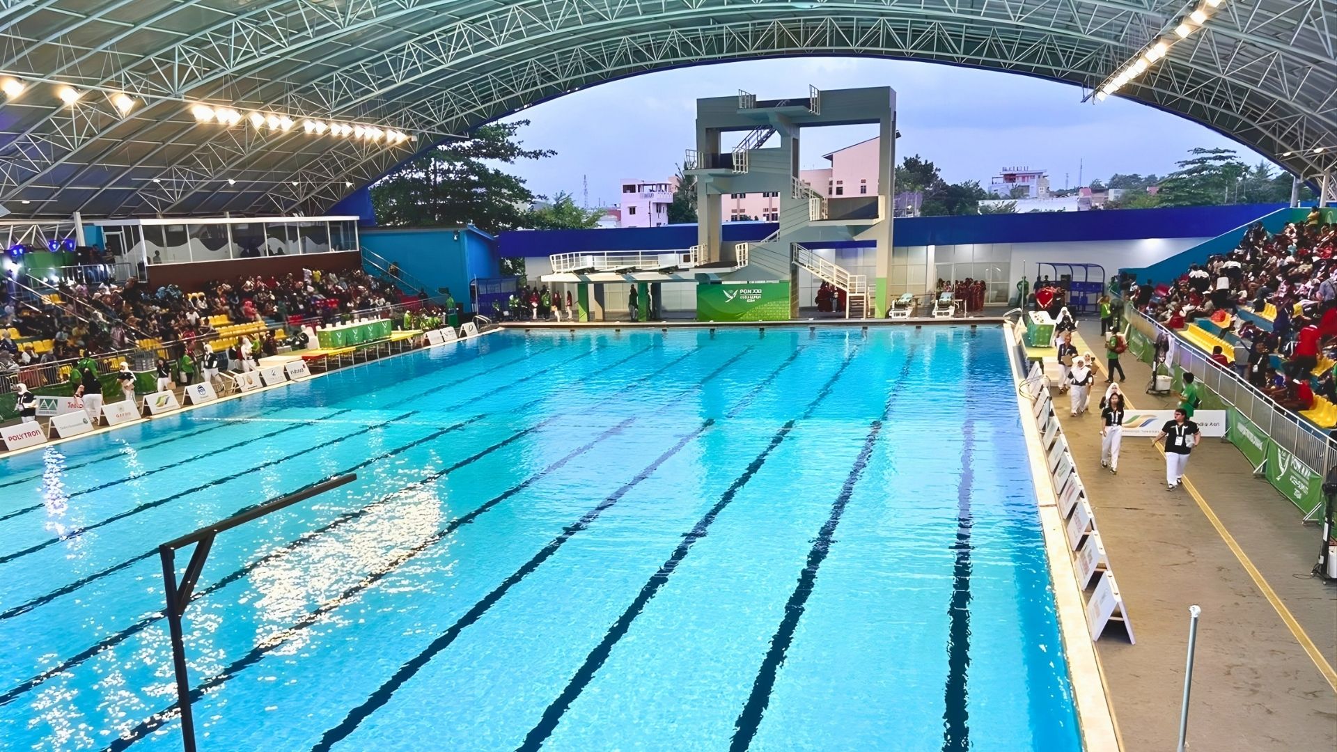 The 50-metre competition pool at the Selayang Aquatic Centre (Kolam Renang Selayang) in Medan, North Sumatra. Spotlights hang from the roof, and light up the indoor area. The spectator stands are filled with spectators.