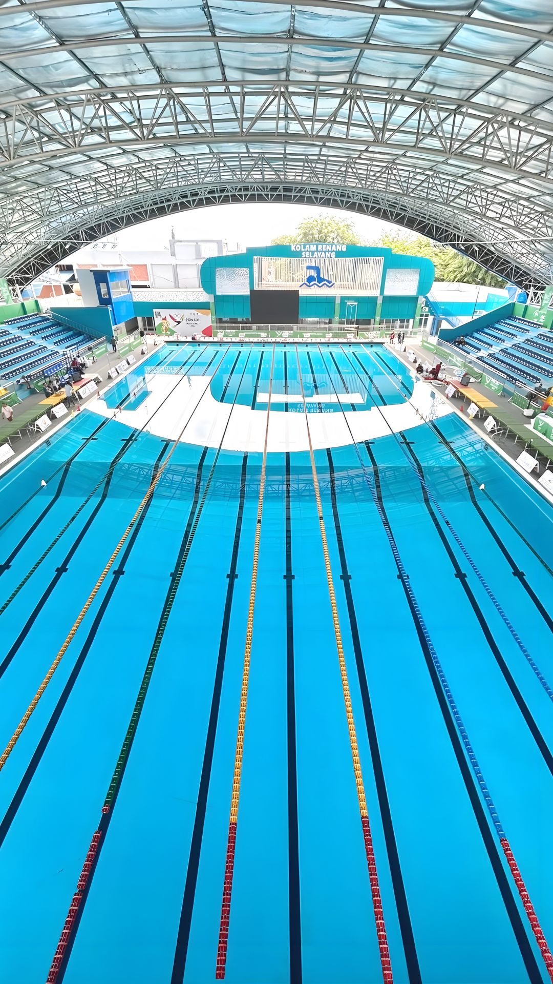 An aerial overview of the Selayang Aquatic Centre. The large competition swimming pool stands out, divided neatly into lanes with pool ropes. Spectator stands line the lengths of the pool, and a signage with “Kolam Renang Selayang” juts out behind.