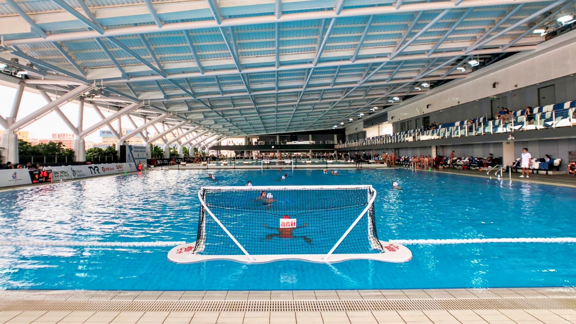 A water polo game is underway at the indoor pool at Our Tampines Hub; a goalkeeper in a red water polo cap stands guard at the opening of the goal post, with other players in the pool.