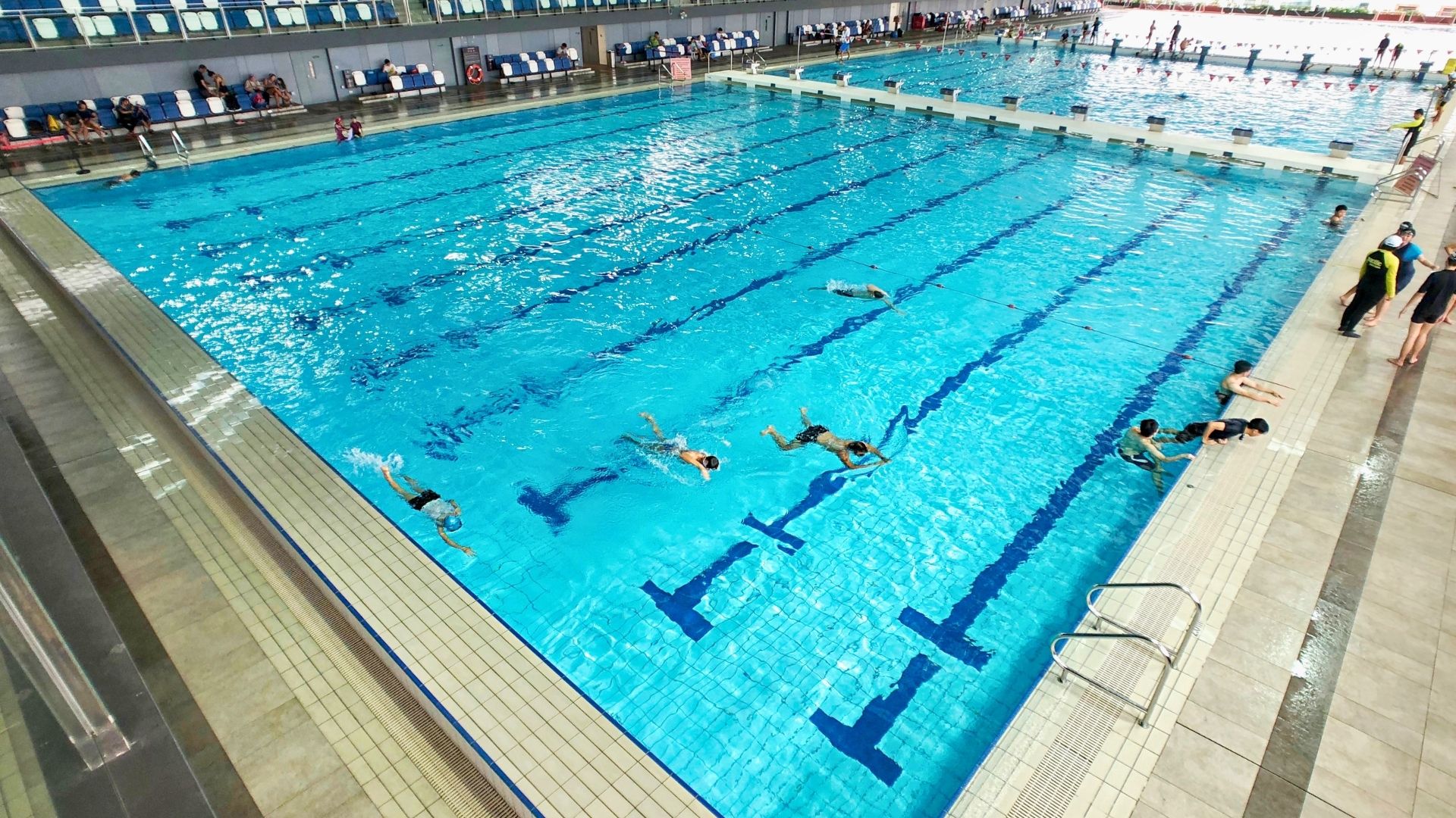 The competition and training pools at Our Tampines Hub Swimming Complex, located indoors with spectator stands along the length. A bulkhead separates the two pools. Swimmers are swimming across the breadth of the pool.